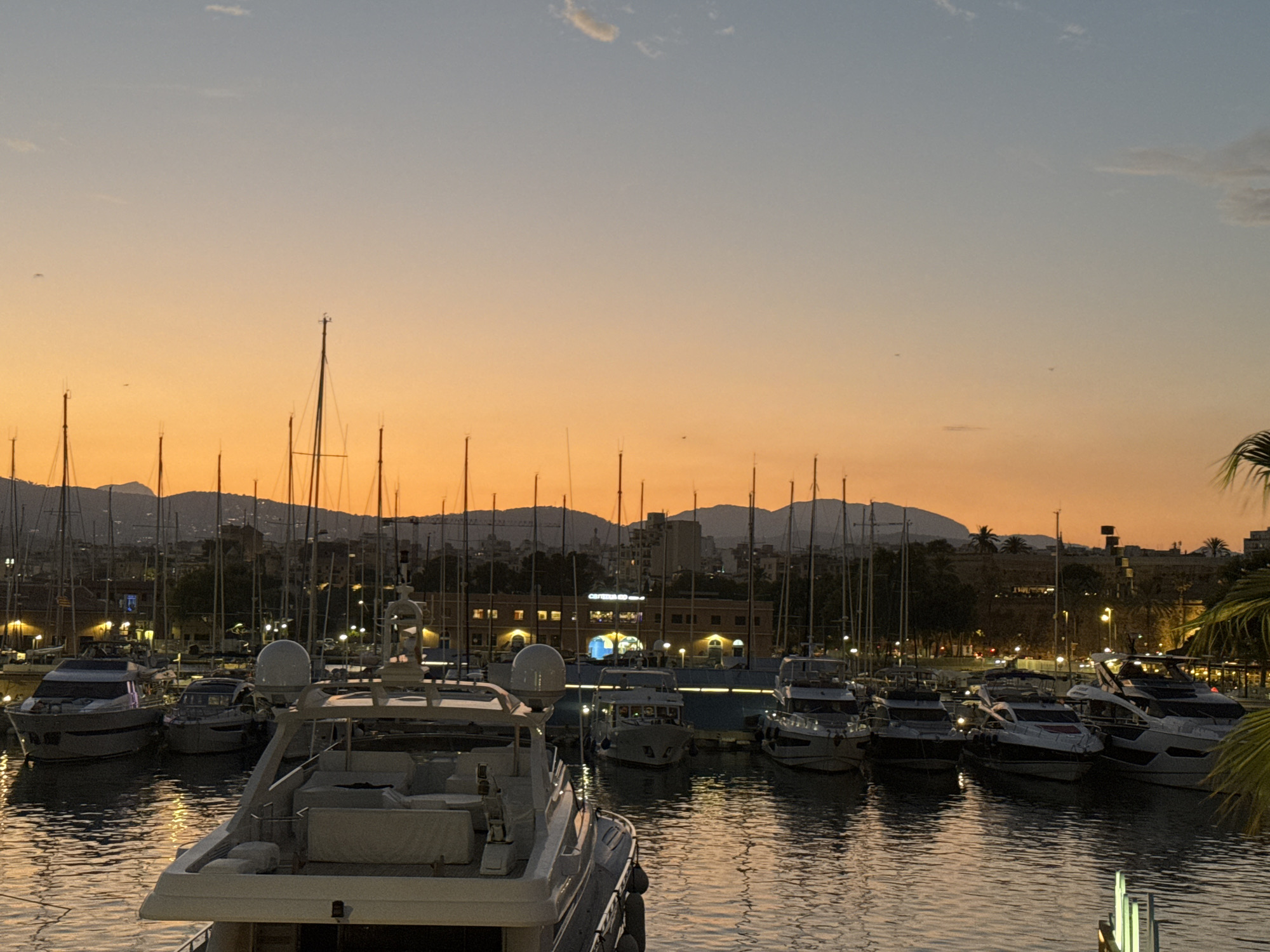 The image depicts a harbor in Palma, Spain, at dusk. Numerous yachts and boats are moored in the water, their masts forming a dense forest against the vibrant orange and pale blue gradient of the sunset sky. The background shows a cityscape with buildings and hills illuminated by soft lights, suggesting the transition from day to night. The water reflects the colors of the sky and the lights from the boats and buildings, creating a serene and picturesque scene. There are no people visible in the image.