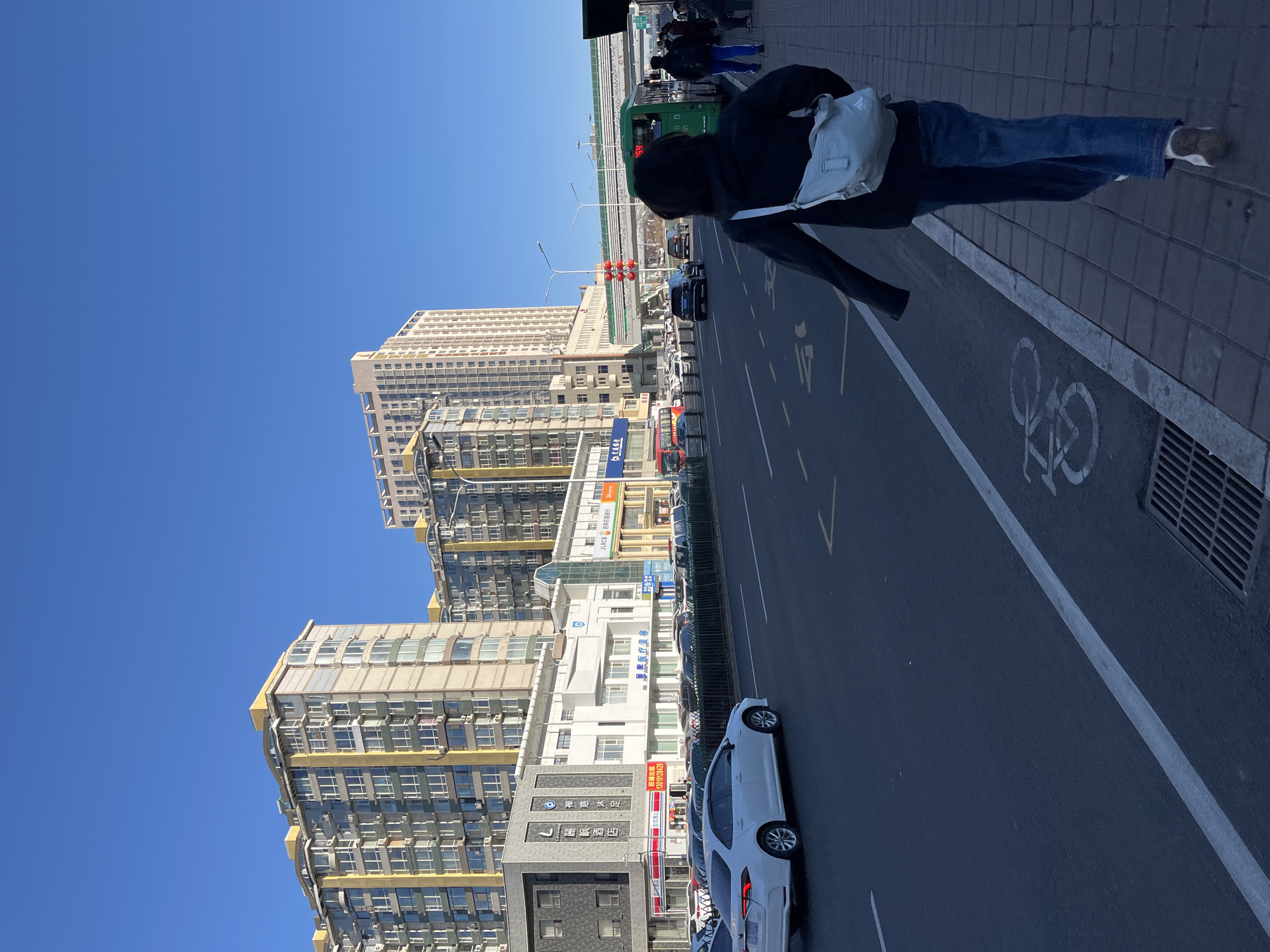 A street scene in Changchun City, China, is depicted under a bright blue sky. The viewpoint is from an elevated position, overlooking the roadside. A pedestrian, seen from the back, walks along the sidewalk, wearing a dark jacket and blue jeans with a white bag over their shoulder. The sidewalk is marked with a white bicycle symbol. The road has several white cars parked alongside, and painted directional arrows are visible on the asphalt. Tall buildings line the left side of the street, featuring both white and yellow facades with numerous windows. In the distance, red traffic lights are visible above a bridge.