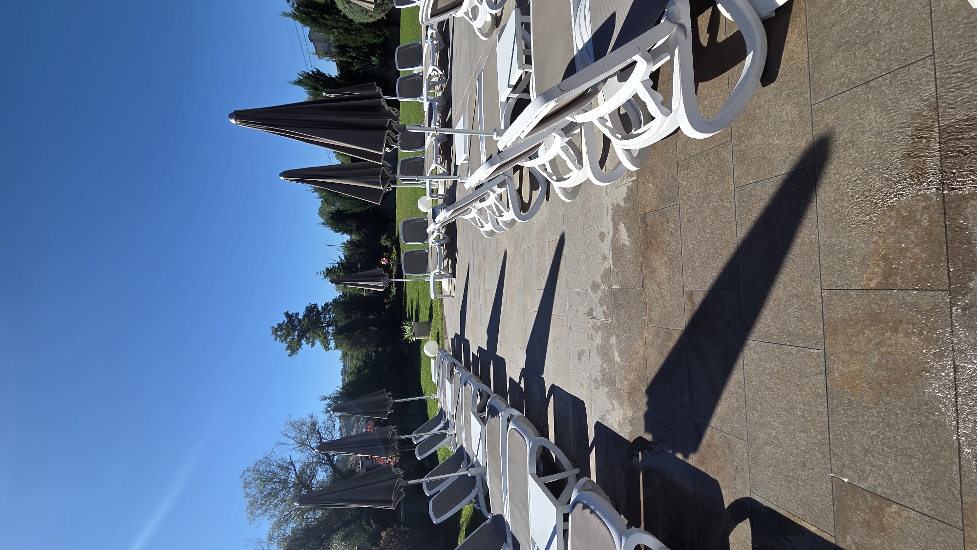 The image shows an outdoor scene with rows of empty white lounge chairs and closed grey umbrellas, arranged on a paved area. The paving stones are large and have a textured, brownish-grey appearance, with some areas appearing wet and reflective. In the background, there is a lush green lawn with trees and bushes. The sky is clear and bright blue, indicating a sunny day. The lighting suggests it is likely morning or late afternoon, given the long shadows cast by the lounge chairs. The overall impression is one of a well-maintained, resort-like setting, possibly a poolside area or a garden patio, ready for use but currently unoccupied.