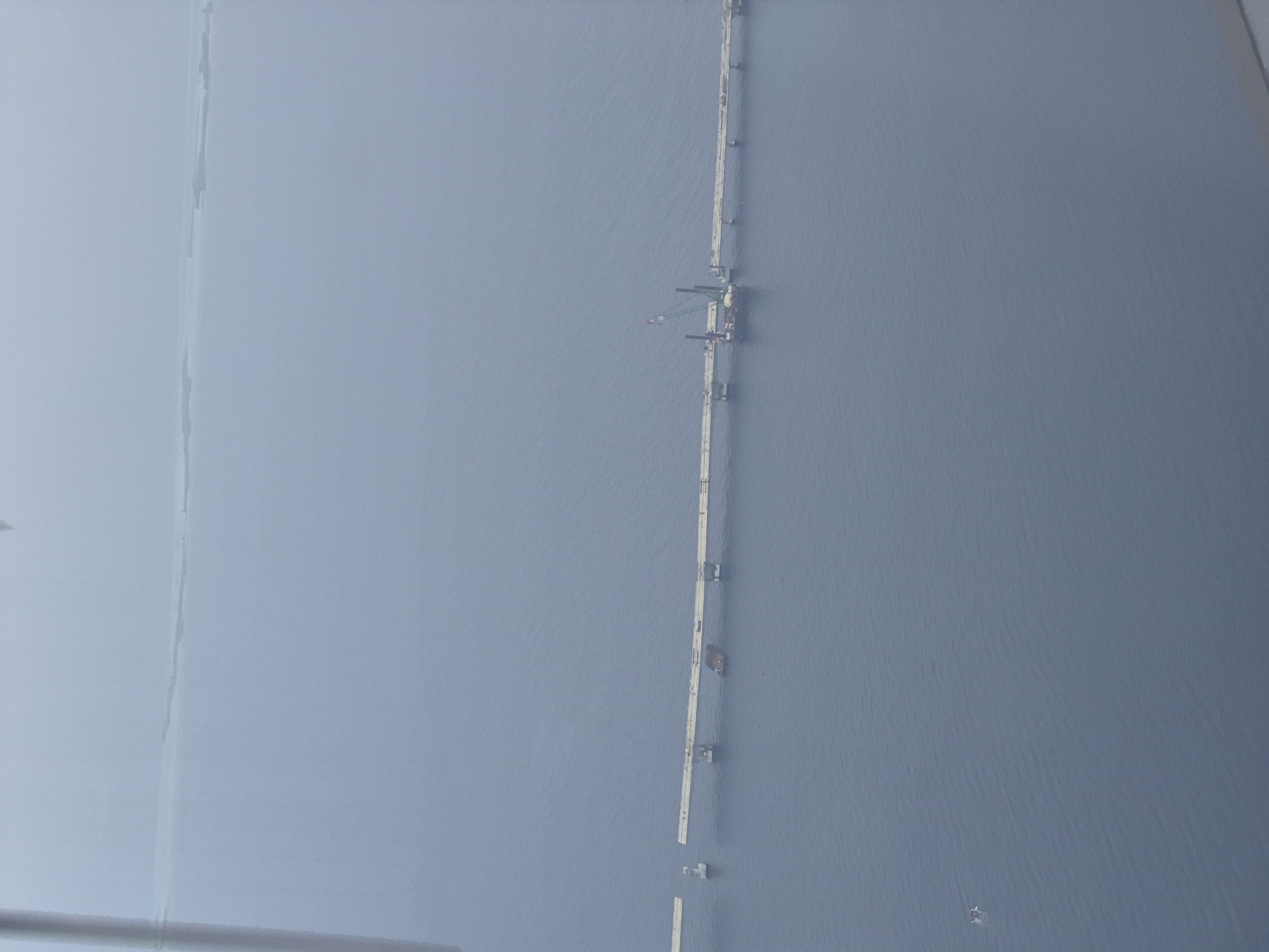 The image shows a long, thin, white structure running vertically through the center of the frame, appearing to be a pier or walkway over water. The structure is composed of multiple segments, some of which are broken or partially submerged. There is construction equipment, including a crane, on one of the intact sections. The water is a uniform gray-blue color. The image is taken from an elevated viewpoint. The left side of the image is obscured by a light gray haze or fog. The location is suggested to be the city of Malé, Maldives.