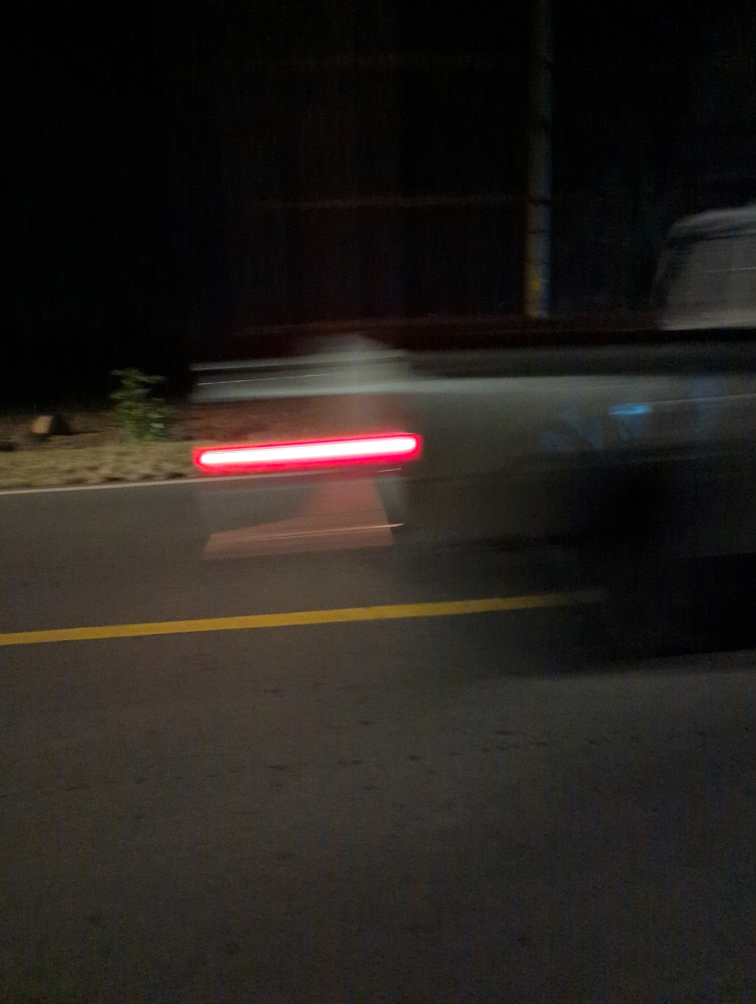 The image depicts a nighttime scene on a paved road in Musanze District, Rwanda. A vehicle's red taillight is blurred, indicating motion. The road has a yellow line marking its edge. The background is dark, with some indistinct shapes that suggest foliage and a vertical structure, possibly a utility pole. The overall impression is one of movement and low light conditions.