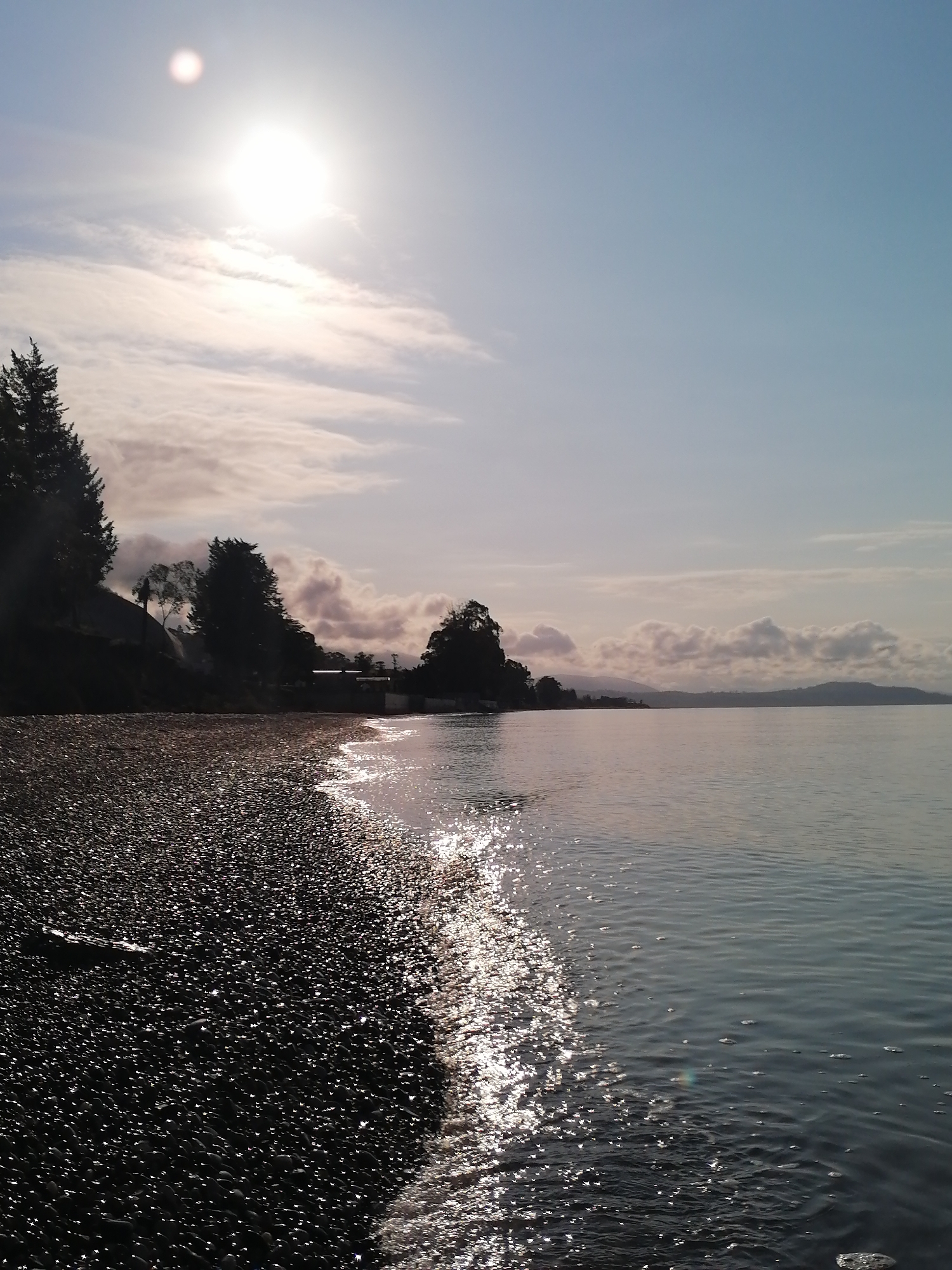 This image captures a serene waterside scene, likely a lake or a calm sea, under a bright, low-lying sun. The foreground features a dark pebble beach on the left, giving way to calm, reflective water on the right. Sunlight glares intensely off the wet pebbles and the water's surface, creating a sparkling, almost shimmering effect.
On the left, a hillside is covered with tall, dark evergreen trees and other vegetation, largely silhouetted against the bright sky and water reflections. Some faint structures or buildings are visible further down the shoreline in the shadows. The sky above is a pale blue, with soft, scattered clouds, particularly concentrated around the intensely bright sun, which appears to be either rising or setting, indicating early morning or late afternoon.
The calm water extends into the mid-ground and background, meeting a distant shoreline characterized by rolling hills or mountains. A line of clouds hovers above these distant landforms, blending with the soft sky. No people or specific events are depicted, contributing to the tranquil atmosphere. The scene evokes a sense of peace and natural beauty. No visible text is present in the image.
