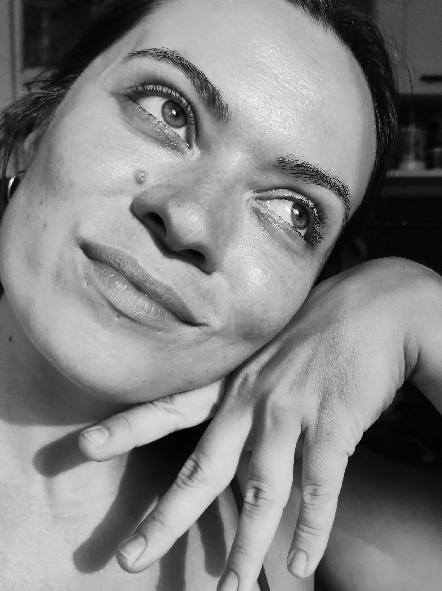 A black and white, close-up, high-angle shot captures a woman's face and hand. She is leaning her chin on her hand, her eyes looking upwards and to the side, with a slight smile on her lips. Her hair is pulled back, and she is wearing a hoop earring. The image is lit from the right, casting shadows on her left side. The background is dark and out of focus. There is no identifiable information about the location in Chaikovsky, Russia.