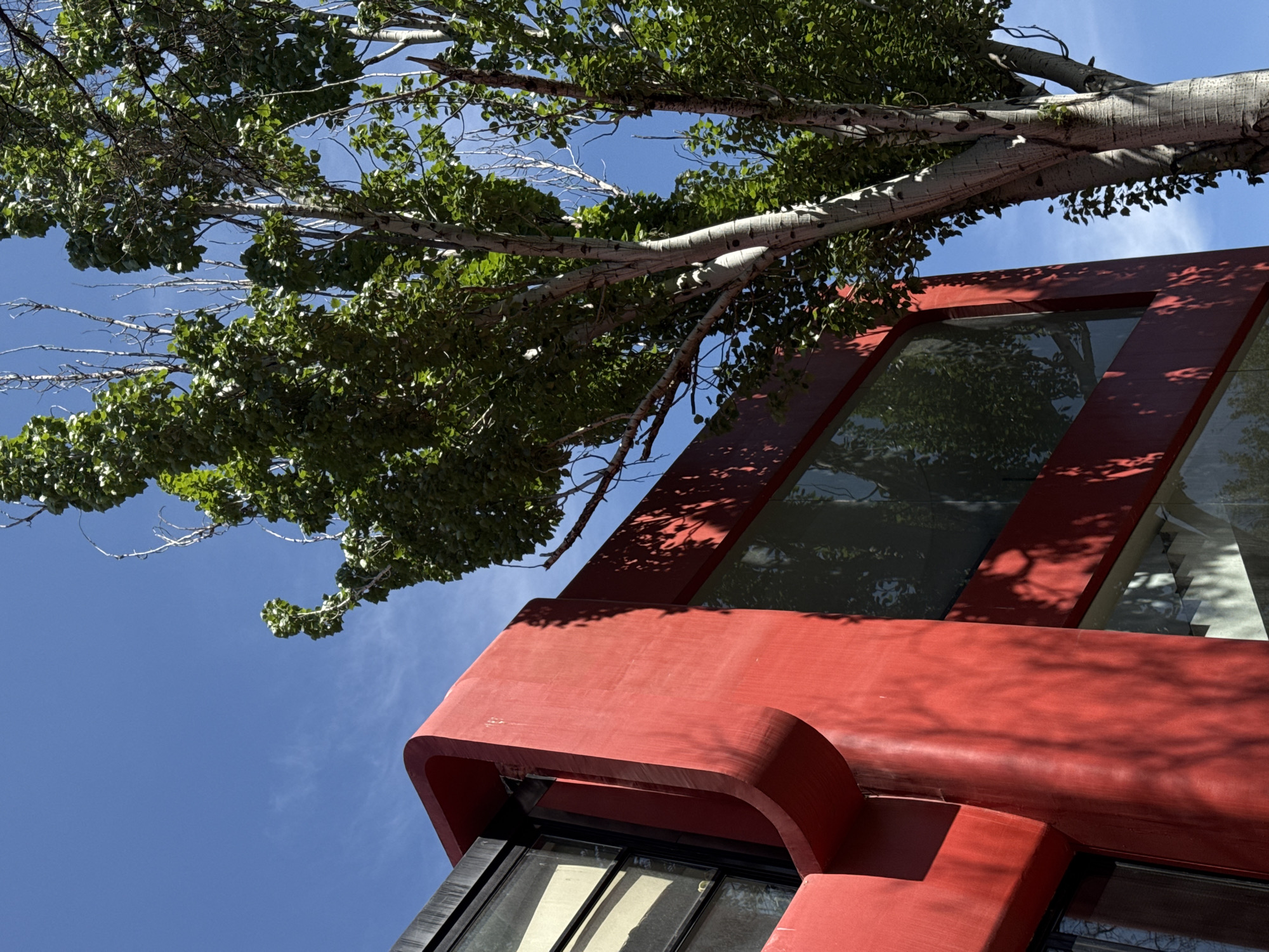 A low-angle view captures a red-painted building under a clear blue sky, partly obscured by a tree with green leaves. The building appears to be modern with large windows reflecting the surrounding environment. Shadows from the tree fall across the red facade. The specific location in Yerevan, Armenia is not discernible from this image. No people, subjects, objects, or actions are evident other than the building's presence and the natural elements of the tree and sky. No interactions are visible.