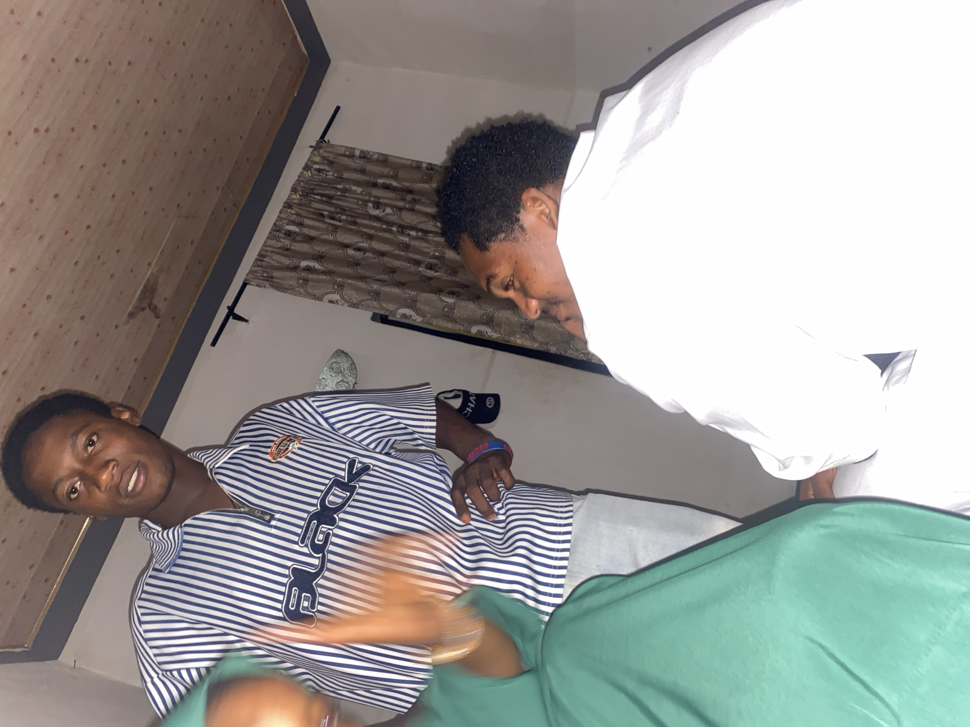 In Bauchi, Nigeria, a group of individuals are gathered in a room with a wood-paneled ceiling. A young man wearing a blue and white striped shirt with the word "BLACK" emblazoned on it stands facing the camera, his mouth slightly open. To his left, a person in a green garment is partially visible, their hands blurred with motion, suggesting they are actively engaged in a gesture or conversation. To the right, another individual, seen from the side, is wearing a white shirt and appears to be looking away from the camera, with their chin resting on their hand. The room's ambiance suggests a casual indoor setting, with patterned curtains visible in the background.