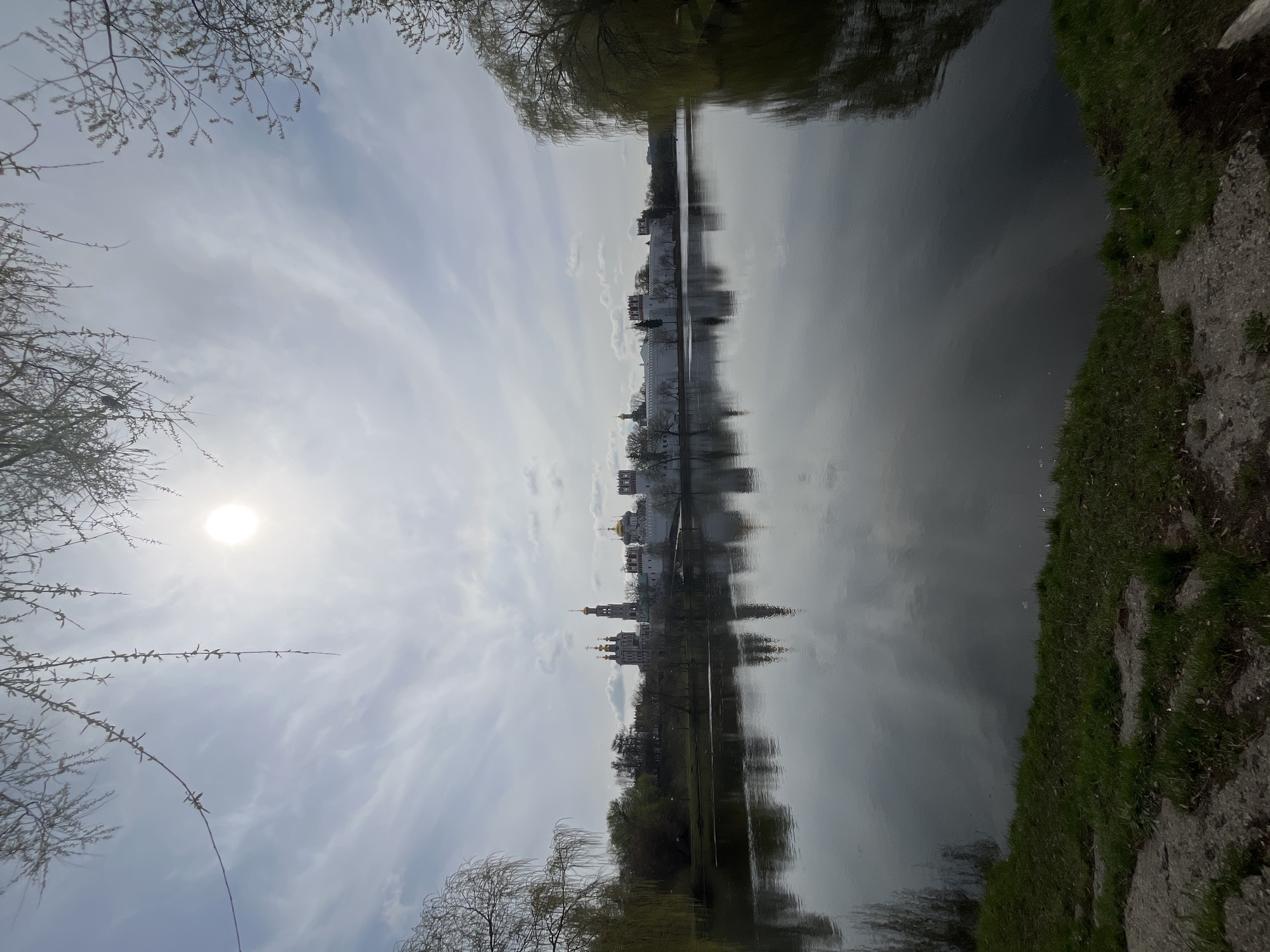 The image shows a body of water reflecting a cityscape, likely a lake or river reflecting buildings. The scene is Moscow, Russia, as indicated by the architectural style of the buildings. The sky is partially cloudy with the sun visible. Trees are present in the foreground, some with bare branches. Vegetation is present on the shore. The composition is such that the reflection is a major element, with the water appearing dark and smooth.