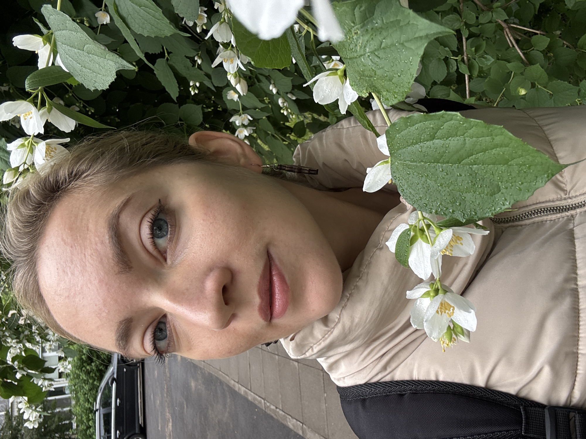 A medium shot selfie shows a blonde woman with blue eyes and fair skin, wearing a beige puffer jacket and a black backpack. She is standing outdoors next to a bush with white jasmine flowers and green leaves, some of which are wet with water droplets. The woman's hair is pulled back, and she is wearing subtle makeup. Behind her, on the left side of the image, is a paved sidewalk and a dark-colored station wagon parked on the street. The scene appears to be in a residential area, possibly in the city of Minsk, Belarus, given the urban environment and vegetation. The lighting suggests it is daytime, likely overcast or after rain.