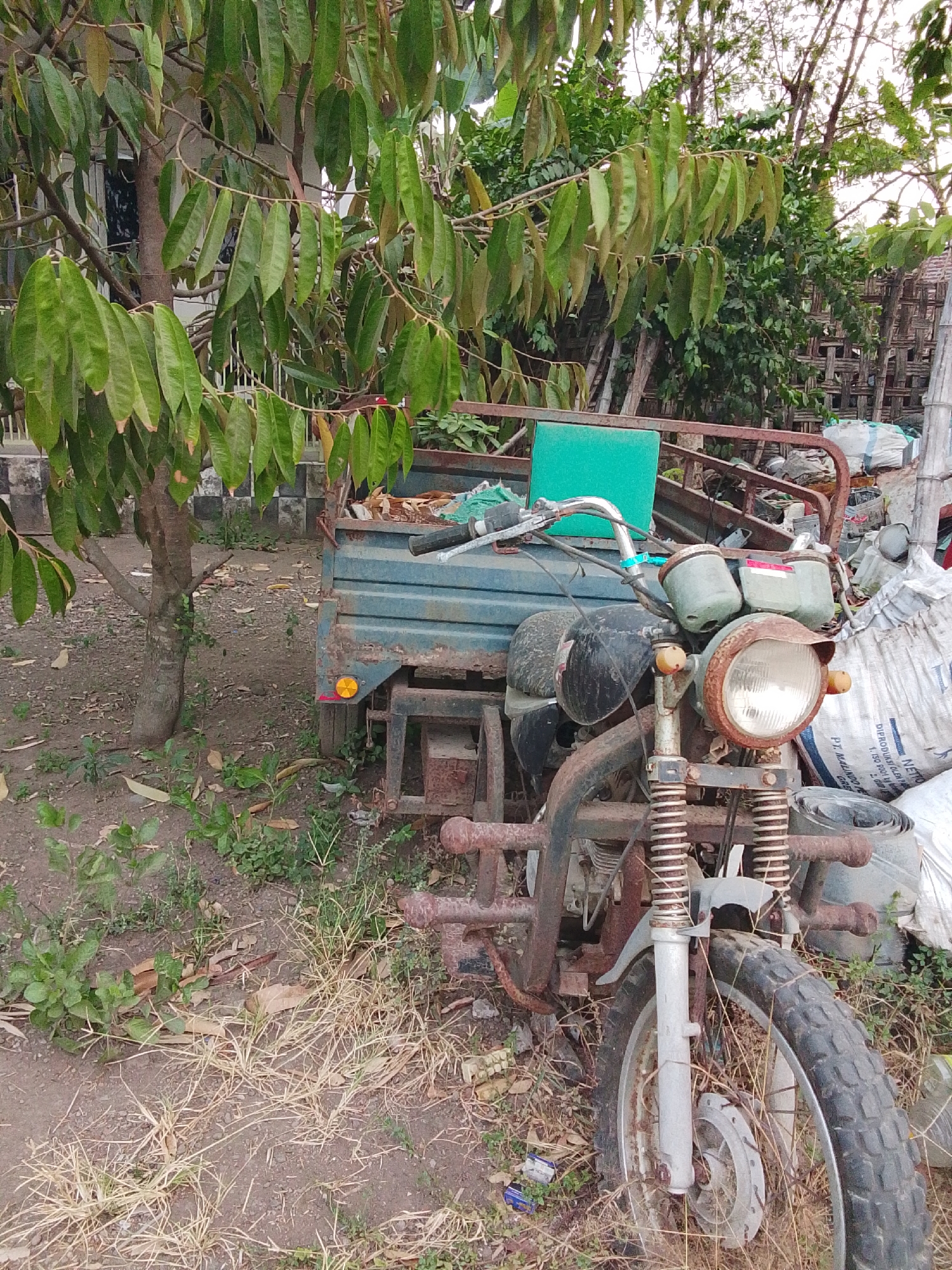 In Bulurejo, Indonesia, an abandoned, rusty three-wheeled motorcycle with a blue cargo bed is parked on a dirt path. The motorcycle is partially covered by the branches of a leafy tree. In the background, a woven bamboo structure and several large white sacks are visible. The ground is covered with dry grass and fallen leaves.