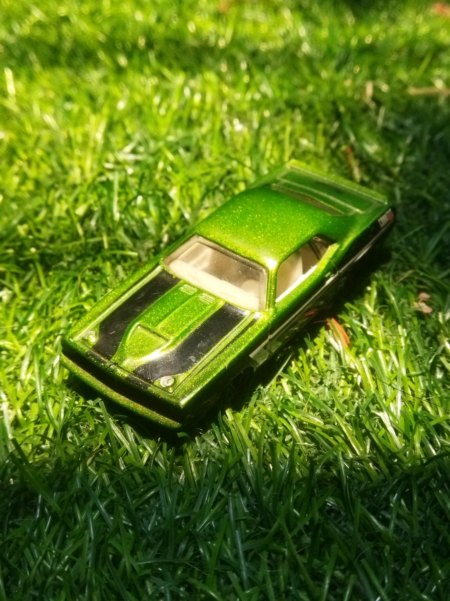 A close-up, eye-level shot shows a bright green toy car with black racing stripes on its hood and roof. The car is positioned on a bed of vibrant green grass, with dappled sunlight casting shadows across the scene. The toy car is the sole focus, with its metallic finish glinting in the light. The background is a blur of green grass, suggesting an outdoor environment. The image was taken in Tarai Bangun, Indonesia.