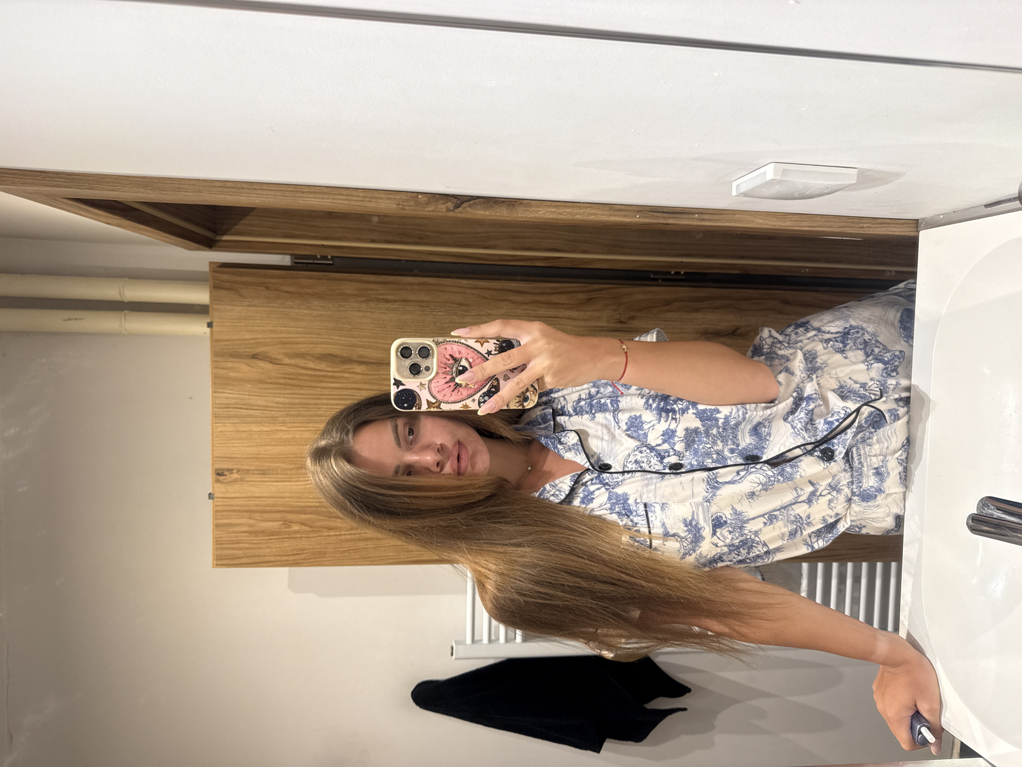 A young woman with long, blonde hair is taking a mirror selfie in a bathroom. She is wearing blue and white patterned pajamas. She is holding her phone in her right hand, which has a decorative case with a pink heart and an eye design. A red string bracelet is visible on her left wrist. Her left arm is resting on the edge of a white sink. To her left, a black towel hangs on a white towel rack. In the background, a wooden cabinet and door are visible, along with a white wall. The image appears to have been taken indoors, likely in a residential setting.