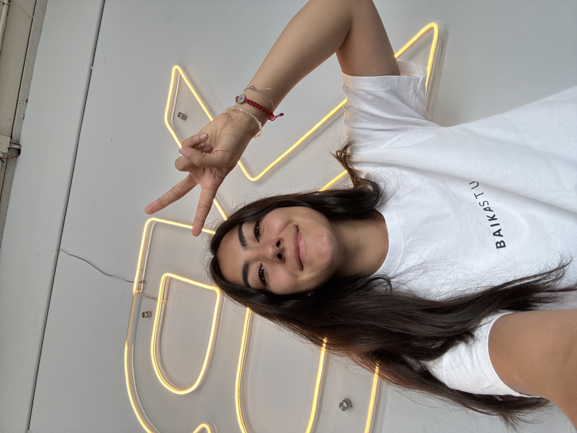 A woman is posing for a selfie indoors, likely in Puebla, Mexico, due to the "BAIKA" text on her white t-shirt, which is a popular brand in the city. She is smiling and holding up a peace sign with her right hand, which is adorned with multiple bracelets. Behind her is a white wall with a yellow neon sign attached. The environment seems to be brightly lit.