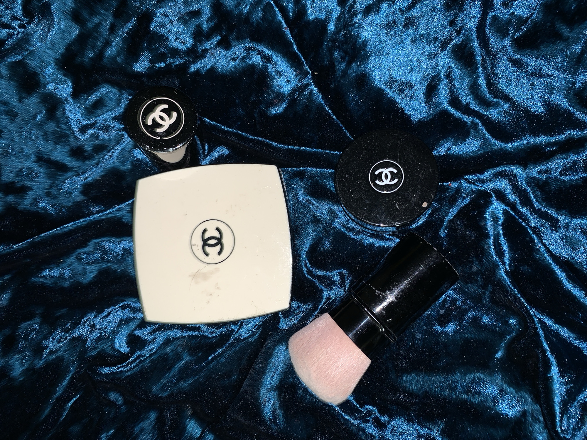 The image shows four Chanel-branded beauty products and a brush on a crumpled dark blue velvet surface. A square compact is located to the left, a round compact is to the right, and a foundation or concealer is positioned at the top left of the image. A brush with a pink head and black handle rests at the bottom right. All items have the Chanel logo on them. The location is unknown.