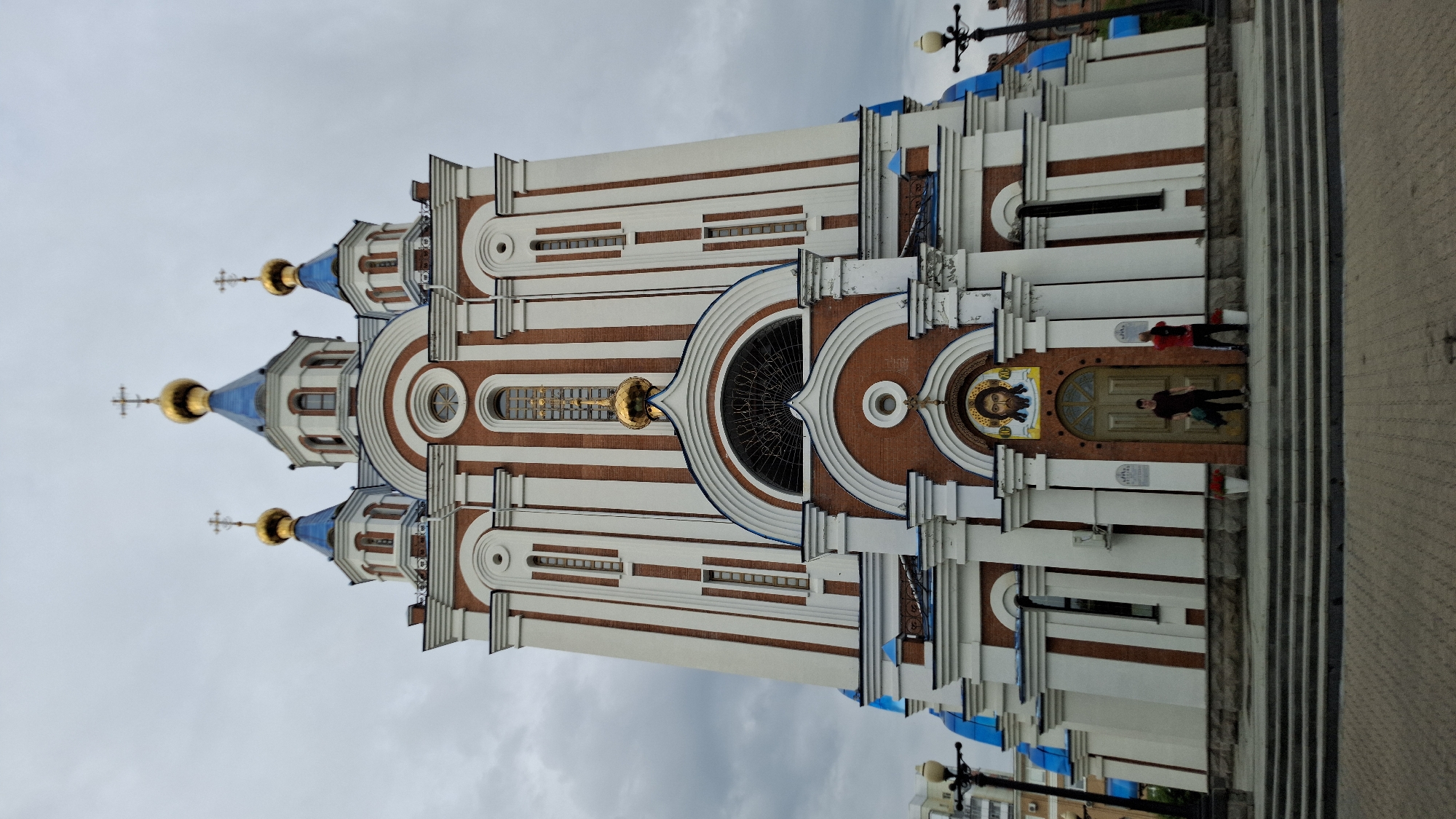 This is a low-angle, full-frame, exterior photograph of the Transfiguration Cathedral in Khabarovsk, Russia. The cathedral is a large, imposing structure with white and red brickwork, topped with blue domes and golden crosses. The front of the cathedral features a prominent entrance with a large wooden door, an icon of Christ above it, and two people standing on the steps. The sky is overcast and gray.