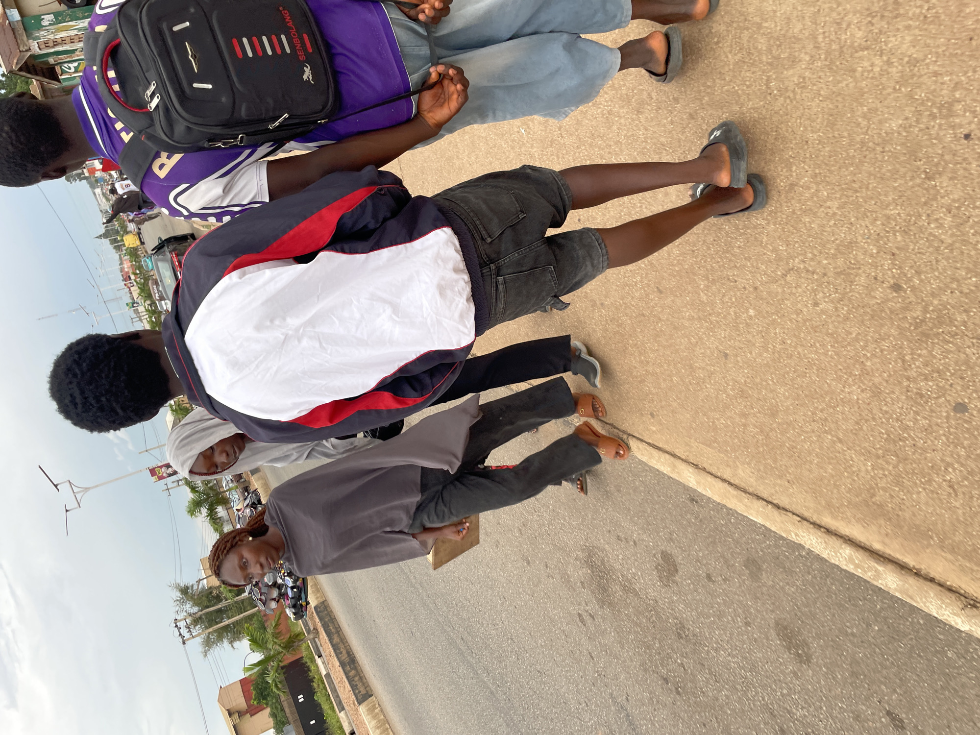 In a street scene in Jos, Nigeria, four individuals are walking on a sidewalk alongside a road. The foreground features the legs and feet of the individuals. A person on the right wears light blue trousers and dark sandals. Next to them, a person in dark denim shorts and black sandals walks ahead. To the left of them, a person in dark trousers and brown sandals is visible. The furthest person on the left wears a dark gray draped garment and black trousers, holding a light-colored flat object. In the background, cars and buildings line the street under an overcast sky. The environment appears to be an urban setting with paved roads and sidewalks.