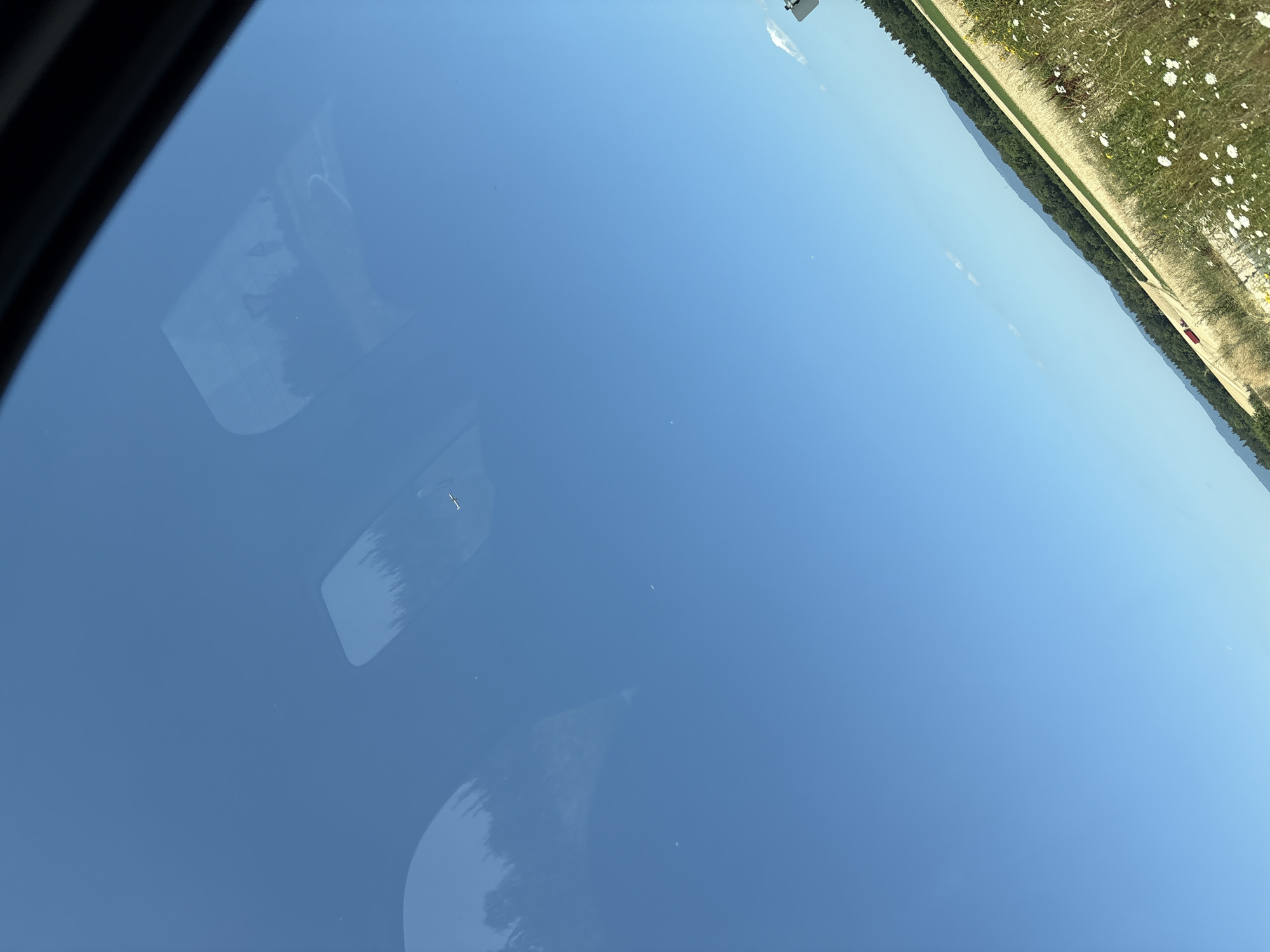 A wide, clear blue sky dominates the upper portion of the image. Reflections of trees and possibly a vehicle are visible on the windshield of a car. In the lower portion, a rural landscape unfolds, with a field of golden crops and a strip of white wildflowers bordering a dirt road. A red tractor is visible in the distance within the field. Hills covered in dark green trees form a backdrop to the field, and a few faint clouds are scattered across the sky. The image appears to be taken from inside a vehicle, looking out through the windshield. The scene is set in Ober-Olm, Germany.