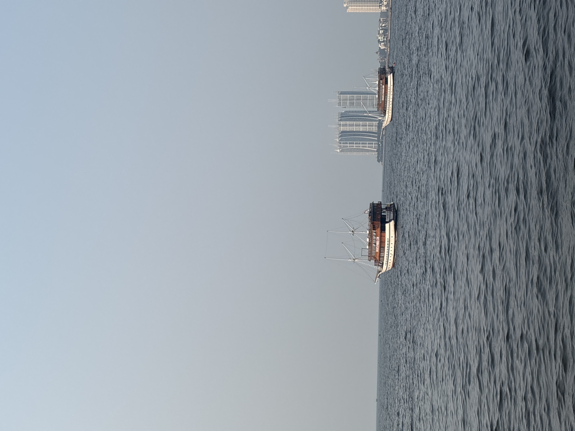 The image depicts a seascape with a hazy sky over the waters of Jakarta Bay, North Jakarta, Indonesia. In the foreground, the dark blue water exhibits moderate ripples. Two traditional wooden boats with white hulls and brown upper decks are sailing on the water. The boat on the left is closer to the viewer and is partially cut off by the frame. The boat on the right is further away and more fully visible.
In the background, a cluster of modern, tall buildings rises from the coastline. These buildings, including a distinctive set of three curved skyscrapers, suggest an urban waterfront. The atmosphere appears slightly overcast or polluted, as indicated by the diffused light and the faint visibility of distant structures. The scene captures a blend of traditional maritime activity and contemporary urban development against a hazy horizon.