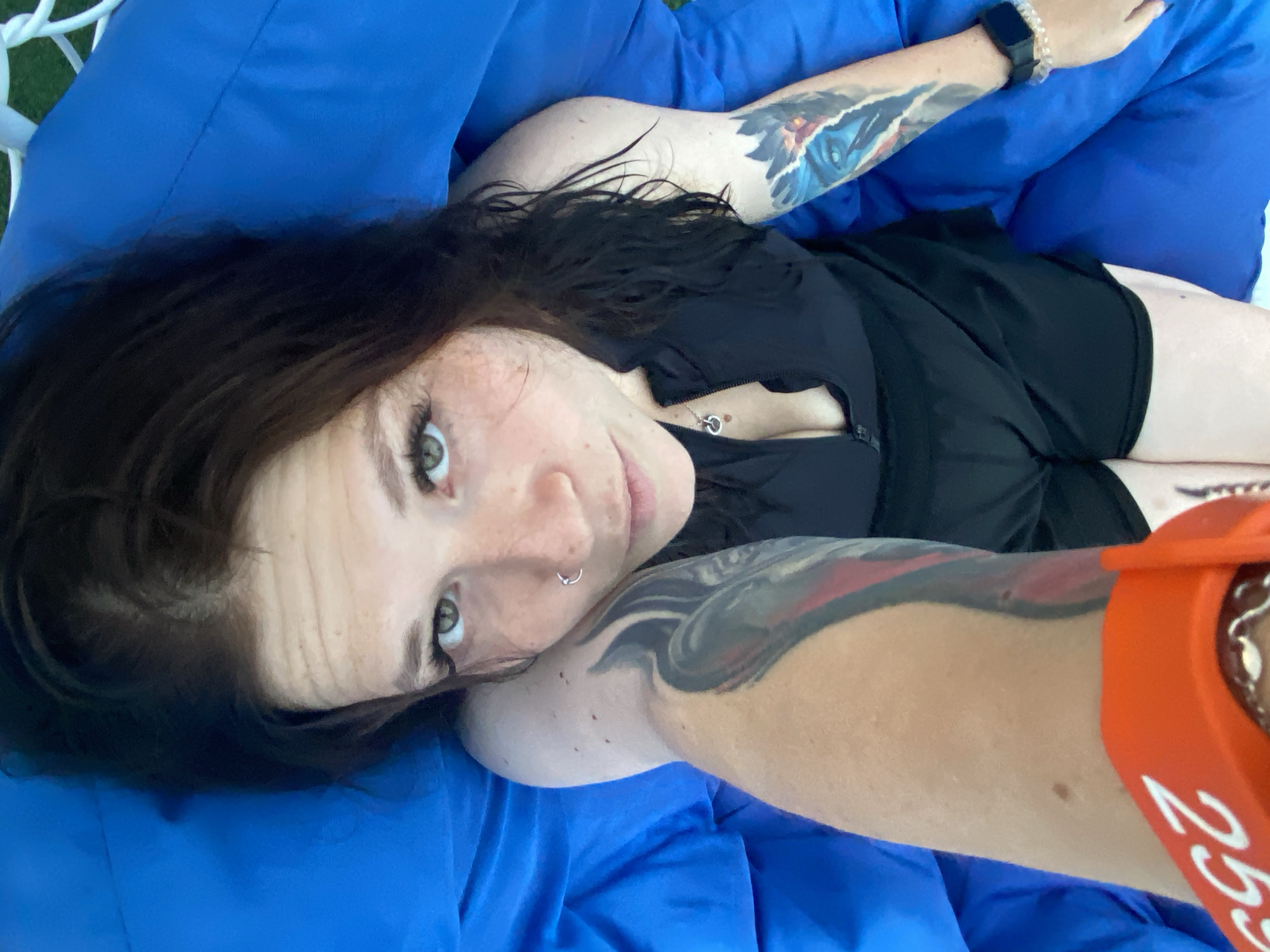 A person with dark hair and green eyes is lying down on a blue surface, possibly a beanbag chair or a hammock. They are wearing a black tank top and black shorts. The person has tattoos on their arms, including one on the left arm with red and black designs and another on the right arm with blue and black designs depicting an eye. They are also wearing a silver nose ring, a silver necklace with a heart pendant, a smartwatch on their right wrist, and an orange bracelet on their left arm with white numbers. The background is out of focus but appears to be outdoors, possibly with some greenery. There is no information in the image to determine its location in Votkinsk, Russia.