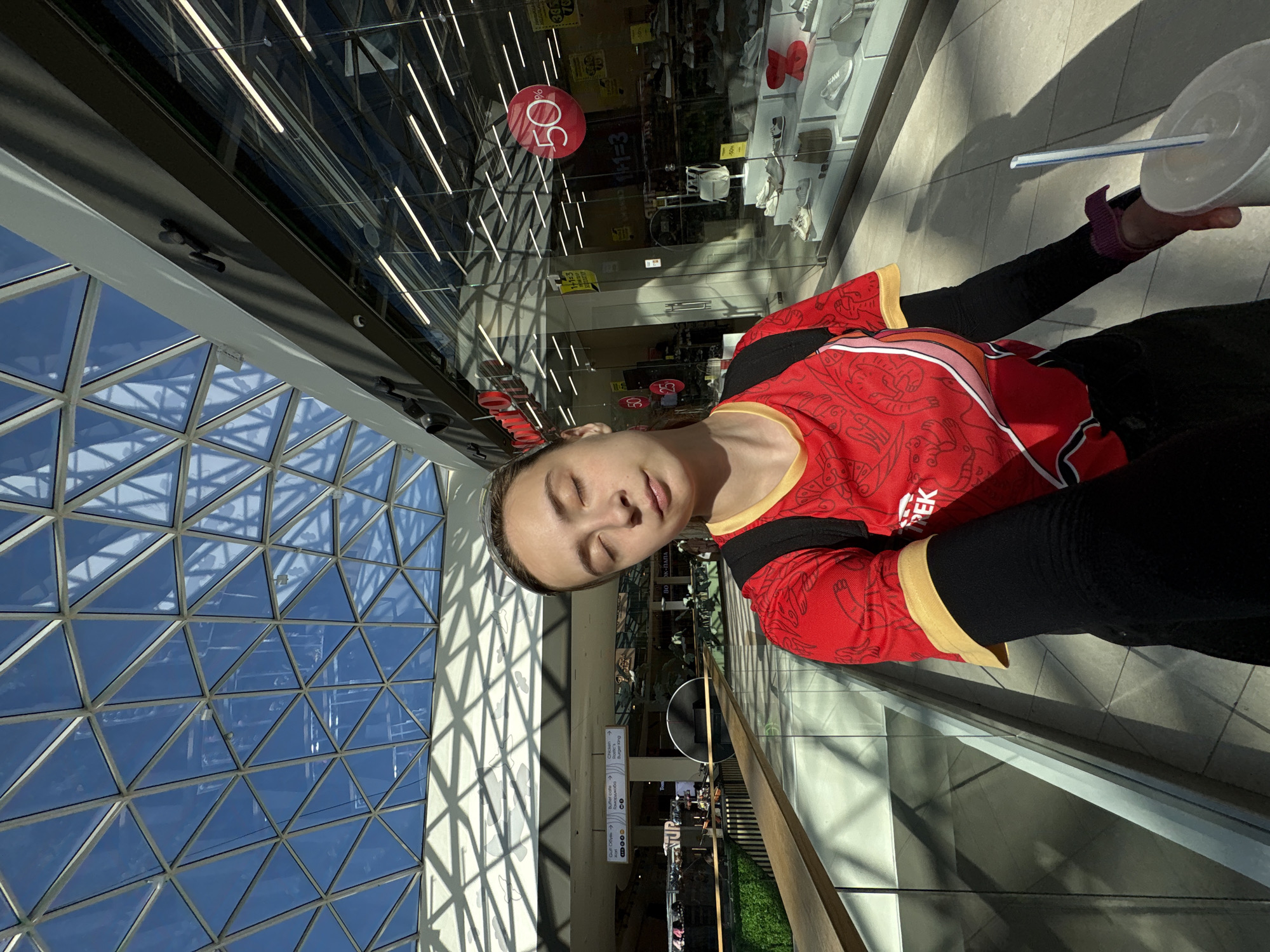 The image captures a woman in a red and black athletic top and black sleeves, holding a white cup with a straw, seemingly in a shopping mall. The woman has her eyes closed. The environment is brightly lit, with a distinctive architectural design, featuring a triangular glass roof structure that allows natural light to flood the interior. Signage indicating "Burger King" and a clothing store advertising a "50%" sale are visible in the background. The sign "Гуф! Обувь" is also visible, which is an advertisement in Russian for shoes. The scene is taking place in the city of Perm, Russia.