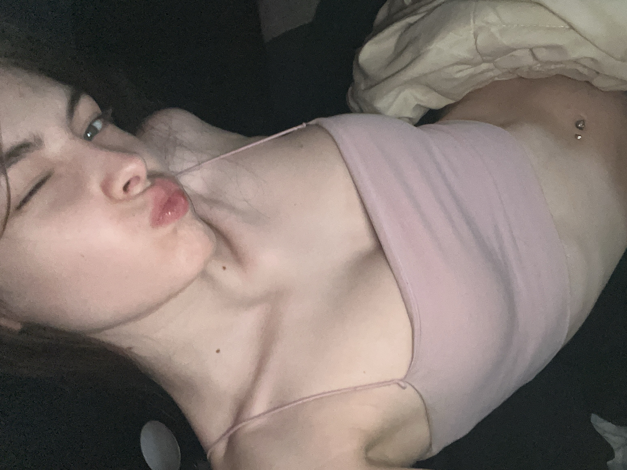 The image shows a close-up, selfie-style shot of a young person in a pink camisole. The person is winking and puckering their lips, with their head tilted to the right. Their hair is pulled back, and a thin strap of the camisole is visible on their shoulder. The background is dark and out of focus, with a hint of a blanket or comforter on the right side. The person has a navel piercing. The image does not contain any elements that can be used to identify a specific location in Sinyavino, Russia, or any other location.