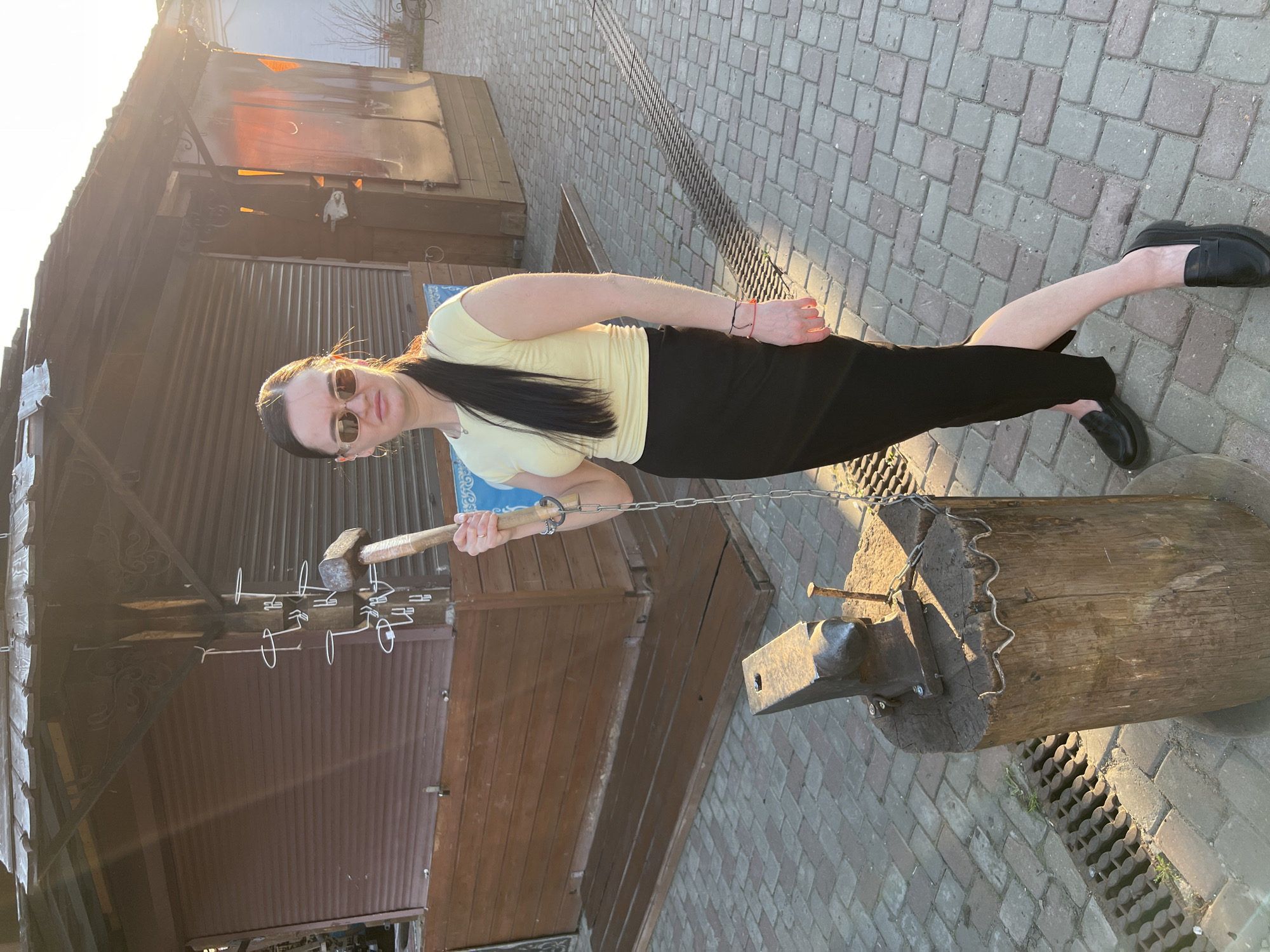 The photograph captures a woman striking a metal object with a hammer near an anvil, outdoors in what appears to be Sviyazhsk, Russia. The woman wears sunglasses, a light yellow shirt, and a black skirt, along with black shoes. She's standing near an anvil mounted on a wooden stump. A metal chain is connected to the stump. Behind her is a structure of wood and metal. The ground is paved with bricks. The lighting suggests the scene is shot either early or late in the day, possibly at sunset.