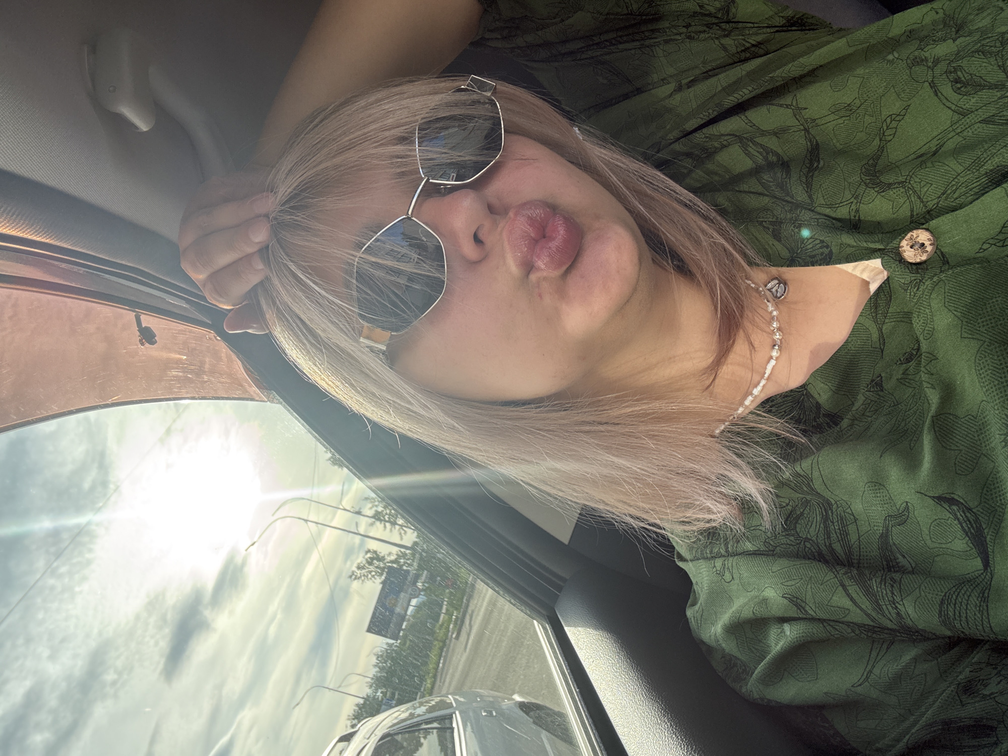 A young woman with blonde hair and sunglasses is taking a selfie from inside a car. She is wearing a green patterned shirt and a pearl necklace. Her left hand is resting on the roof of the car, and her right hand is touching her hair. The sun is shining brightly through the car window, creating lens flares. The background shows a street with cars and trees, suggesting an outdoor setting in a city. The image could have been taken in Birobidzhan, Russia, given the presence of streetlights and billboards common in urban areas of the region.
