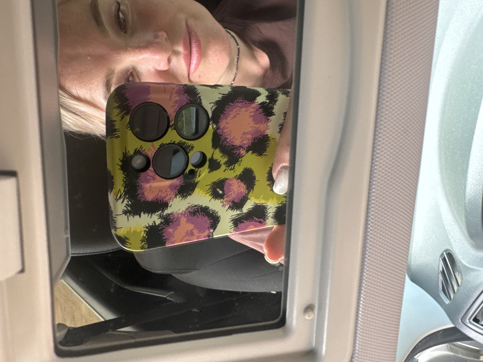 The media file shows a reflection of a person's face and a cell phone in a mirror. The person, who appears to be female with blonde hair and light-colored skin, is looking down towards the phone. Her lips are slightly parted and painted with a pinkish-red lipstick. She is wearing a dark-colored top and a thin necklace. The cell phone has a brightly colored leopard print case with shades of yellow, pink, and black. The phone's camera array is visible, featuring multiple circular lenses.
The reflection is captured within a frame that suggests the interior of a vehicle, possibly a car, given the visible textures of what appears to be a car interior. The lighting in the scene is moderate, indicating it's likely daytime, though the exact time of day is not discernible. There are no explicit weather cues.
The setting appears to be indoors or within a covered space, as evidenced by the reflections and the framing of the mirror. The overall mood is casual, with the subject capturing a selfie or documenting her surroundings. No text is visible in the media file. The location context provided is Nozhovka, Russia, but there are no specific visual cues within the image to confirm or deny this.