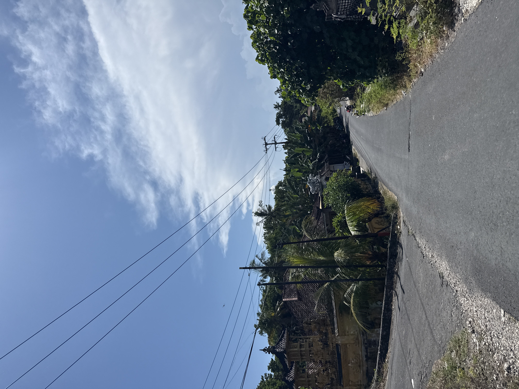 The image shows a narrow, paved road winding uphill in Batununggal, Indonesia. Power lines run along the left side of the road against a blue sky with scattered white clouds. Lush green tropical vegetation and buildings line both sides of the road. A person is visible further up the road, walking uphill.