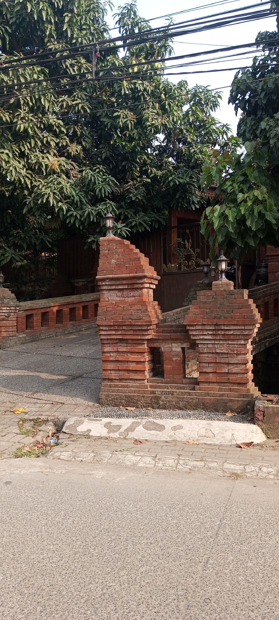 The image shows an outdoor scene with a brick gate structure and trees. The gate is made of red bricks and has two lamps on top of it. There is a stone pathway leading up to the gate. Behind the gate, there are lush green trees with dense foliage. The ground in front of the gate is paved with stones and asphalt. There are some leaves scattered on the ground. In the background, a building with wooden elements and decorative metalwork can be seen. The sky is visible at the top of the image. The scene appears to be a public space or entrance to a property in Setu, Indonesia, possibly a residential area or a place of business, given the presence of mature trees and the style of architecture.