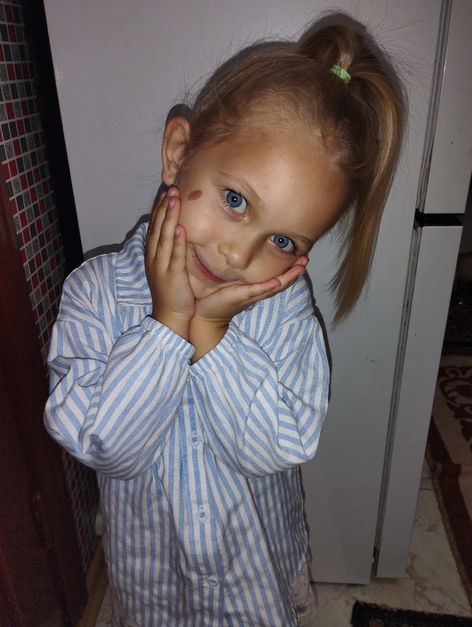 In Fălești, Moldova, a young blonde girl with bright blue eyes is photographed in a playful pose. She is wearing a blue and white striped pajama top and has her hands pressed against her cheeks, a slight smile on her face. A small brown birthmark is visible on her left cheek. Her hair is pulled back in a high ponytail, secured with a green hair tie. The background consists of a plain white wall and a glimpse of a tiled surface to the left. The lighting suggests an indoor setting.