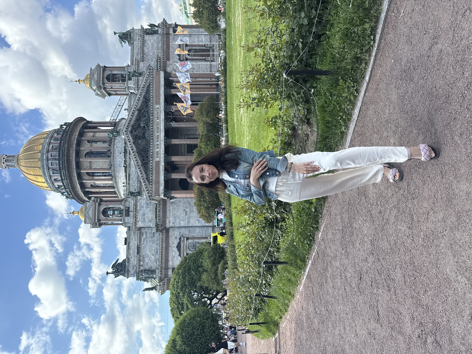A woman stands in front of St. Isaac's Cathedral in Saint Petersburg, Russia. The woman is wearing a denim jacket, a white t-shirt, and off-white pants. She is also wearing light purple sandals. She is standing on a gravel path, with a manicured lawn and a hedge behind her. In the background, St. Isaac's Cathedral is visible, with its large golden dome and classical architecture. Several flags are flying in front of the cathedral. The sky is blue with white clouds.
