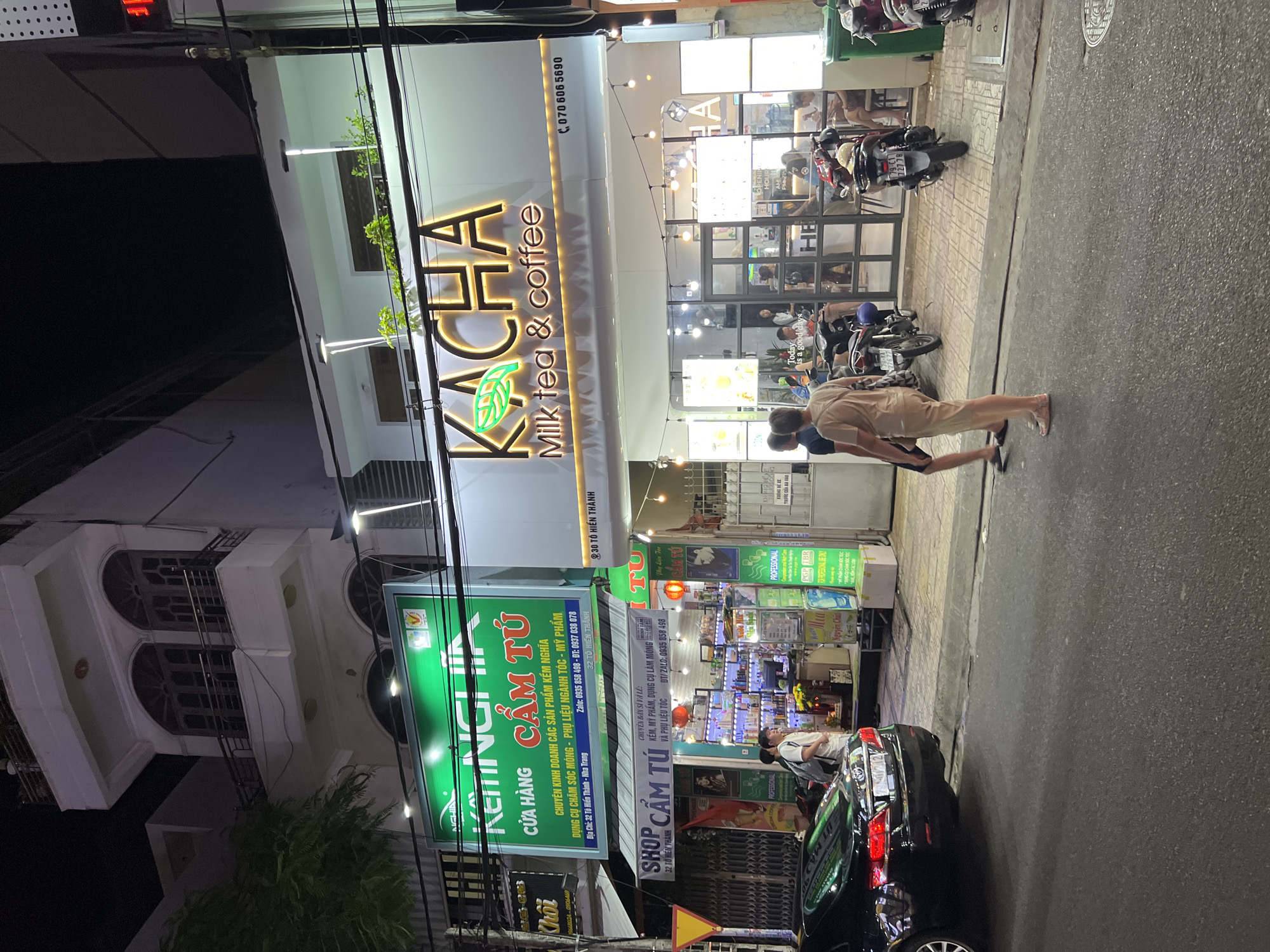 The image shows a street scene at night in Nha Trang, Vietnam. On the right side of the image is a building with a prominent sign that reads "KACHA Milk Tea & coffee". The building has a modern storefront with glass windows and doors, decorated with string lights. Inside, patrons are visible sitting at tables. Several motorcycles are parked in front of the building.
To the left of "KACHA", another storefront is visible with a large green sign displaying text in Vietnamese, appearing to be a shop selling cosmetic and hair products. This sign also includes the address "32 Tô Hiến Thành - Nha Trang". A black car is parked on the street in front of this shop. Two people are standing on the sidewalk near the car. The street appears to be paved, and there are sidewalks. The overall environment suggests a commercial area with shops and businesses open at night.