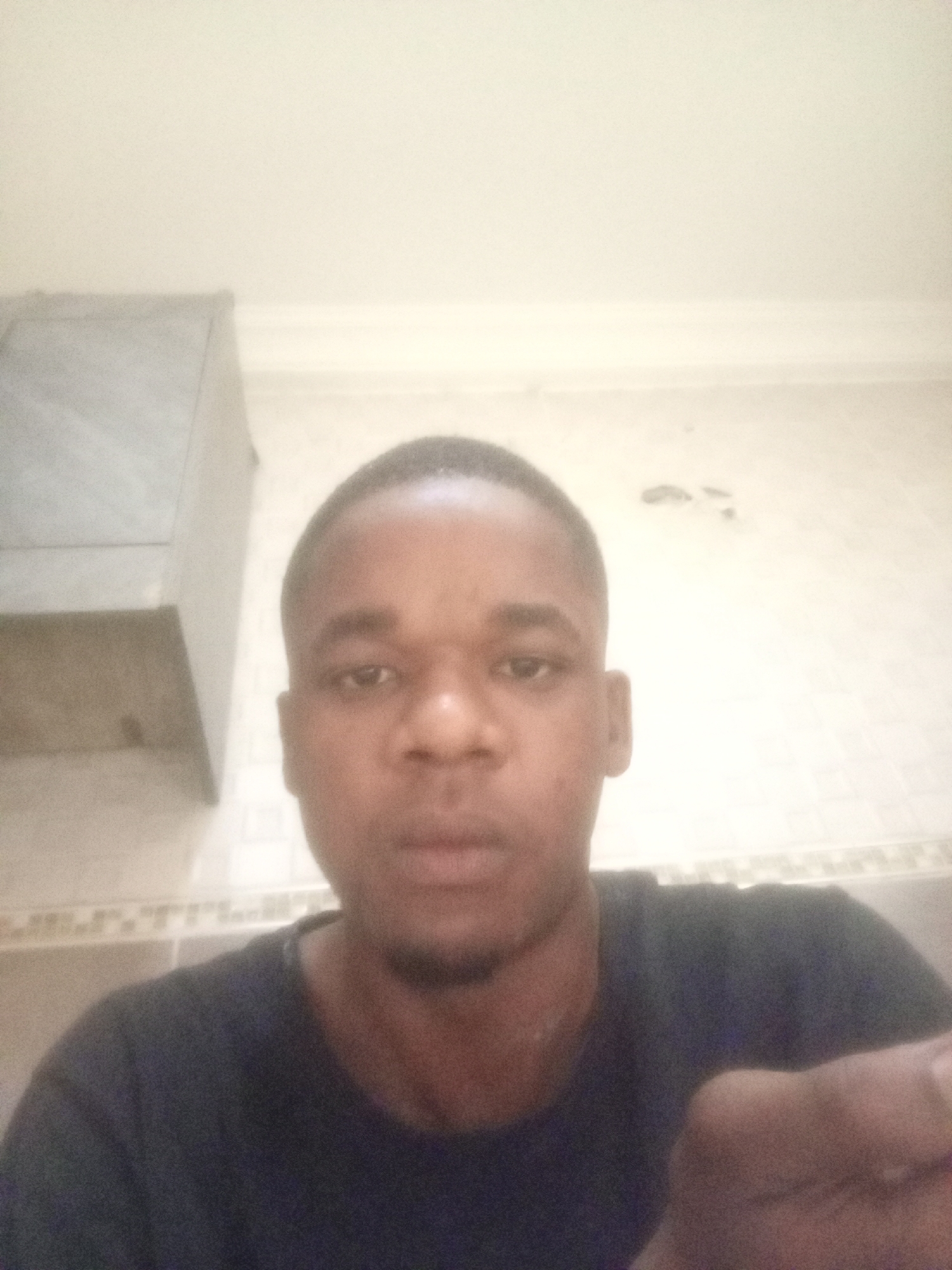 A close-up, slightly blurry selfie of a young Black man with a short, cropped haircut. He has a dark complexion, a rounded nose, and slightly parted lips. He is wearing a dark-colored T-shirt. The background appears to be an interior shot, likely a kitchen or bathroom, indicated by the presence of a cabinet in the upper left corner and tiled trim along the wall. The wall is off-white with some discoloration or patches visible. Considering the prompt, the location is presumed to be somewhere in Kubwa, Nigeria, but there are no specific details in the image that confirm this.