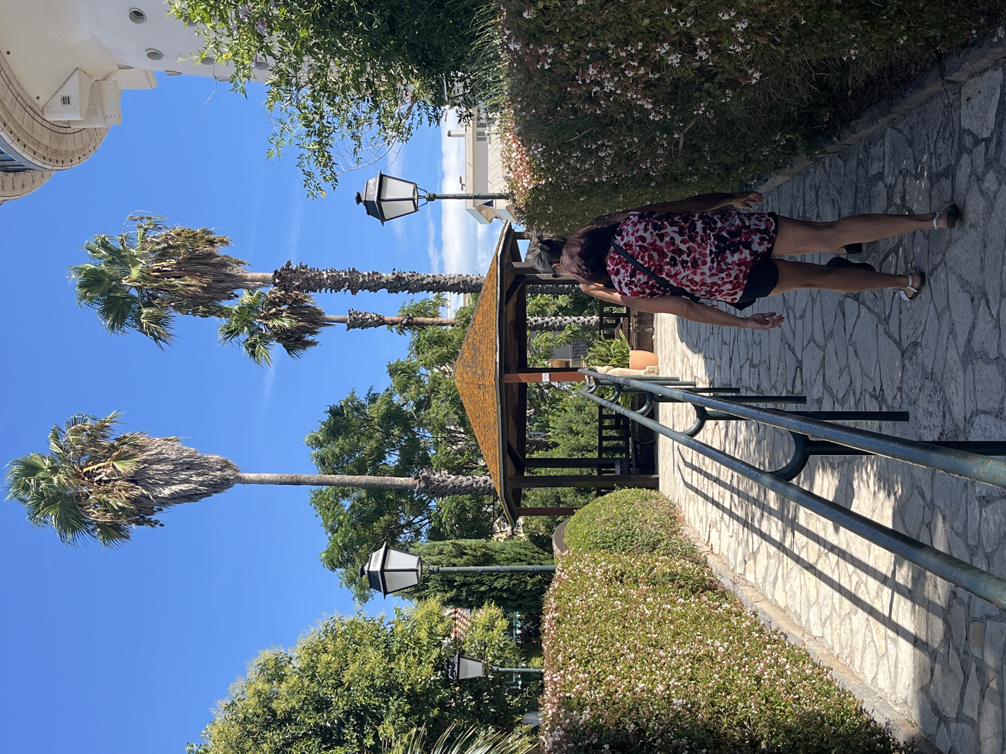 The image depicts a sunlit pathway in Palavas-les-Flots, France, leading towards a wooden gazebo surrounded by lush greenery. The path is paved with irregular flagstones and lined with neatly trimmed hedges dotted with small white flowers. A dark metal railing runs along the right side of the path, casting long, sharp shadows across the flagstones.
In the background, several tall palm trees with dried fronds tower against a clear blue sky. A classic-style street lamp with a black hexagonal lantern stands near the railing. The gazebo, with its orange-brown roof and dark wooden structure, is visible further down the path, partially obscured by foliage. In the far distance, a large, modern white building with circular windows can be seen.
Two individuals are walking away from the camera on the pathway. The person in the foreground is wearing a sleeveless red and white floral print top and black shorts, with sandals on their feet. Another person, dressed in darker clothing, is walking slightly behind them.