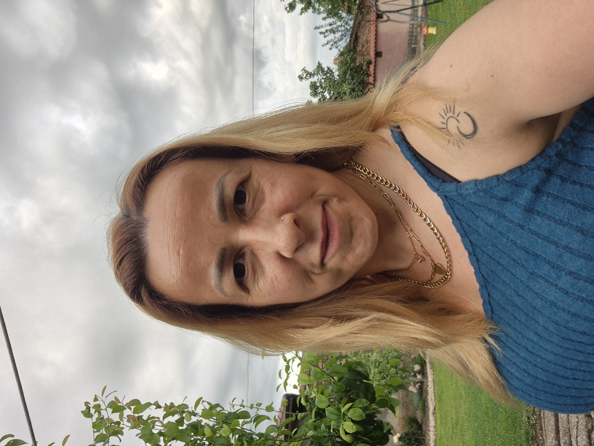 A medium-shot, eye-level, outdoor selfie captures a light-skinned woman with shoulder-length blonde hair and light brown eyes. She is looking directly at the camera with a slight smile. She has a subtle nose ring and a small mole on her left cheek. She is wearing a teal ribbed tank top layered over a black bra strap visible on her left shoulder. Multiple gold chain necklaces are visible around her neck. A small tattoo of a crescent moon and sun is on her upper left arm. The background shows a garden with green grass, a bush with green leaves on the left, and a red-tiled roof structure behind her right shoulder. The sky is overcast. The image is in color and appears to be taken during the daytime.