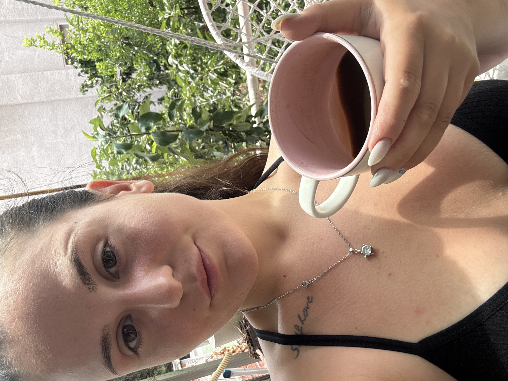 A young woman is taking a selfie outdoors, holding a mug of coffee. She has dark hair pulled back and is wearing a black tank top. A silver necklace with a pendant is visible. There is a tattoo on her left shoulder blade. The background shows greenery and a white hanging chair. The specific location in Plovdiv, Bulgaria cannot be determined from the image.