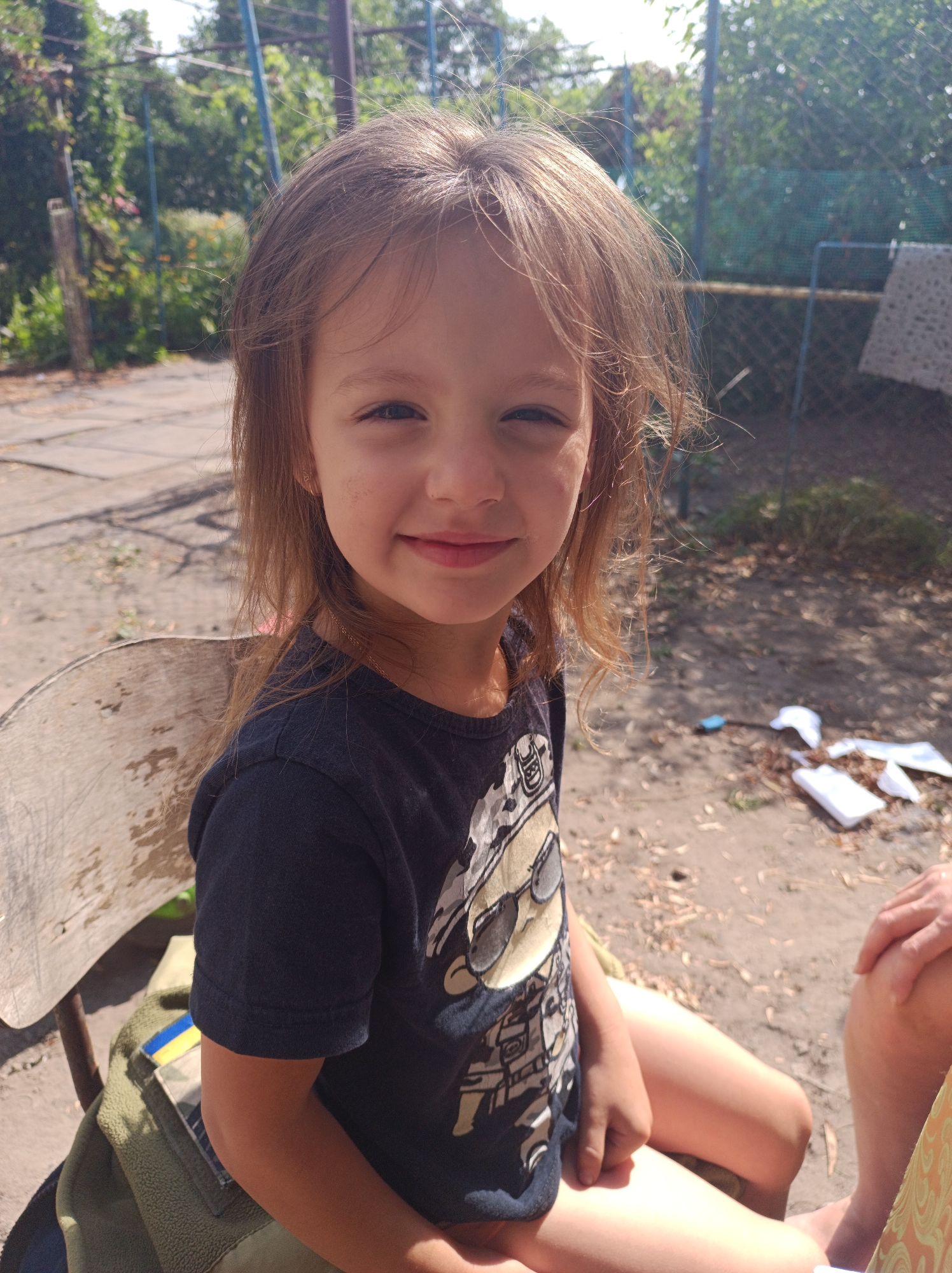 A young girl with long, light brown hair and fair skin is sitting on a wooden chair outdoors. She is looking at the camera with a slight smile, her eyes are dark and her eyebrows are light. She is wearing a dark blue t-shirt with a graphic print on it. Her arms are resting on her lap, and her legs are visible. Behind her, there is a dirt ground with some scattered leaves and twigs. In the background, there is a chain-link fence, some greenery, and a white cloth hanging on the fence. The scene appears to be in a rural or suburban setting, possibly a backyard or a courtyard. The lighting suggests it is daytime with sunlight. On the chair next to the girl, there is a green bag with a patch that resembles the Ukrainian flag. The overall mood of the image is casual and relaxed.