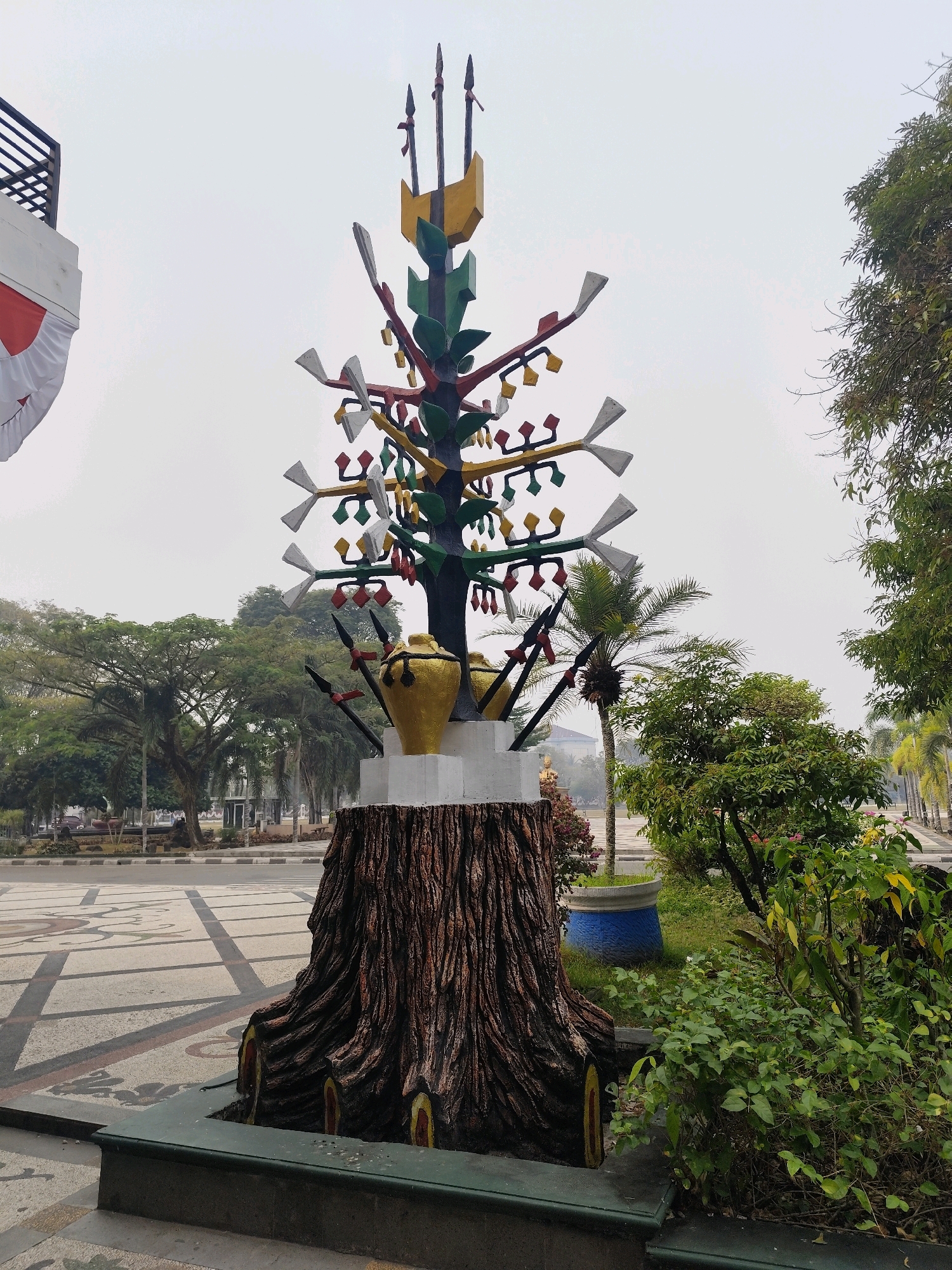 An outdoor, eye-level shot captures a unique, colorful sculpture in Palangka Raya, Indonesia. The sculpture, positioned in the foreground, is a stylized tree with a thick, textured base resembling a tree stump. The stump has visible circular cutouts painted with red and yellow, suggesting cross-sections of a tree trunk. Above the stump, the sculpture branches out into a vibrant tree-like structure painted in green, red, and yellow, adorned with white and yellow leaf-like shapes. Two large, golden urns are prominently placed on either side of the tree's base. The sculpture is surrounded by lush green foliage and some flowering plants.
Behind the sculpture, the environment transitions to a paved public space with intricate patterned tiles. A road with parked cars is visible in the mid-ground. Further back, a line of mature trees forms a backdrop, with some palm trees interspersed among them. In the far distance, buildings are faintly visible through a hazy atmosphere. To the left of the sculpture, a portion of a building with a black railing and a white facade draped with a red and white banner, likely the Indonesian flag, is present. The sky is overcast, contributing to the diffused lighting of the scene. The overall impression is that of a cultural or civic landmark in a public park or square.
