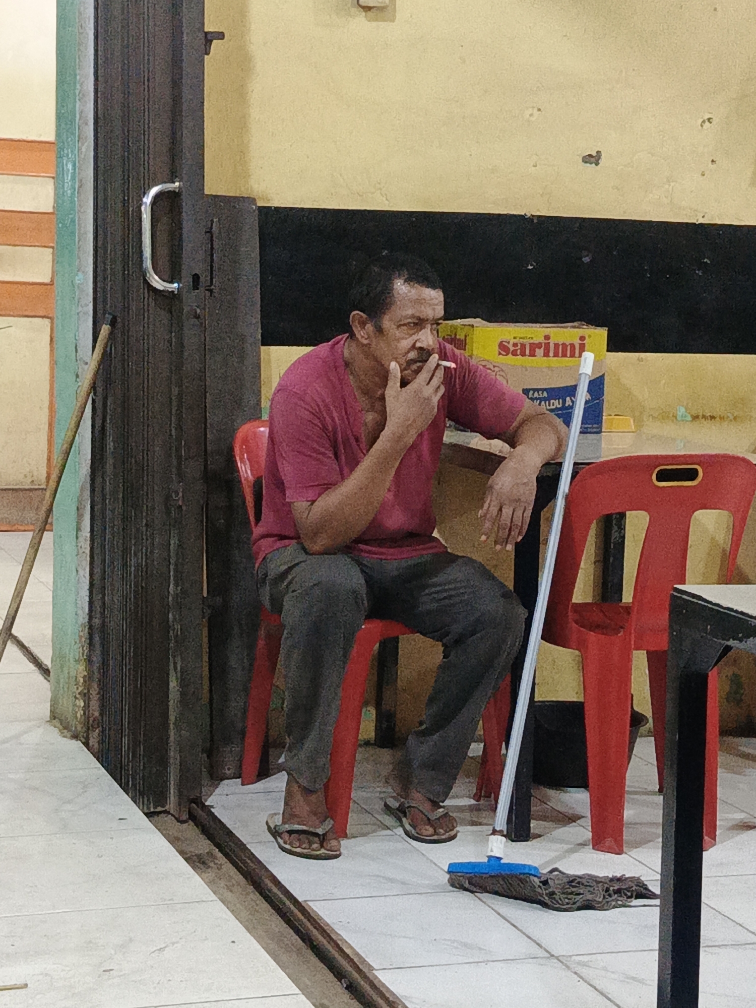 In Sangso, Indonesia, a single adult male is depicted seated on a red plastic chair within what appears to be a modest establishment. The man, with dark hair and a mustache, wears a maroon t-shirt, dark grey trousers, and brown flip-flops. He holds a lit cigarette in his right hand, positioned near his chin, while his left forearm rests on a dark-colored table.
To the man's immediate left, a dark, corrugated or slatted metal folding door or barrier, equipped with a chrome pull handle, is partially visible. Further left, a wooden stick or handle leans against a wall. The background features light yellow walls with a horizontal black stripe. Behind the man, a yellow cardboard box labeled "sarimi" and displaying "RASA KALDU AYAM" is visible.
On the tiled floor to the man's right, a blue and white mop handle with a dark, stringy mop head lies. A black bucket is partially visible beneath the table. Another unoccupied red plastic chair is positioned to the right of the table. The overall scene suggests a break or resting period in a functional, possibly commercial, environment.