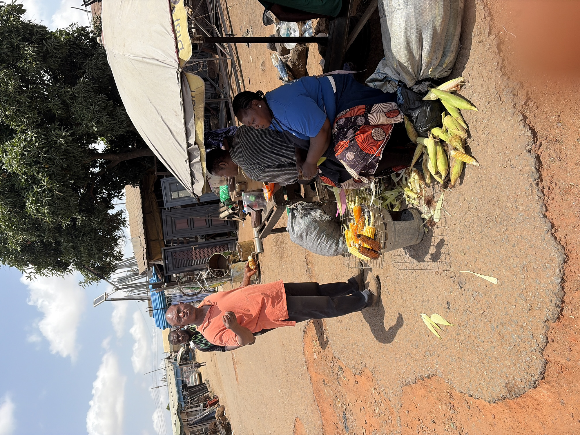 The image depicts a roadside scene in Gura Top, Nigeria. Two women are seated under a makeshift canopy, sorting through a pile of fresh corn, some still in their husks, alongside a large sack. A man in an orange shirt and dark pants stands nearby, holding a piece of corn. A second man is partially visible behind him. The background includes buildings, trees, and a partly cloudy sky. The road is asphalt and shows signs of wear. There are metallic objects visible in the background. The scene appears to be a local market or roadside vendor selling fresh produce.