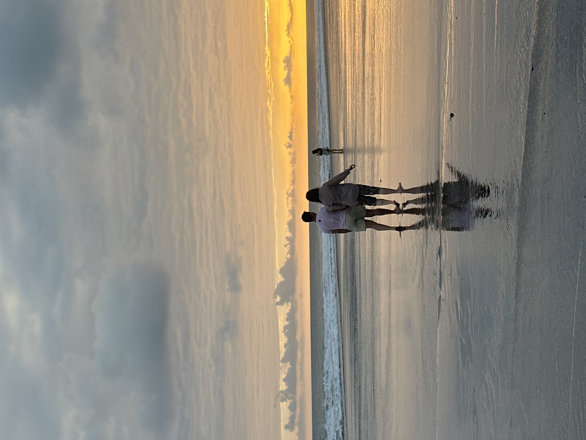 A couple walks hand-in-hand on a beach in North Kuta, Indonesia, during sunset. The sky is a blend of pale blue and warm orange hues, with streaks of clouds creating texture. The ocean, with gentle waves, reflects the warm light of the setting sun. The wet sand of the beach also mirrors the sky, creating a glossy surface. In the distance, a lone figure stands in the shallow water, partially obscured by the waves. The couple in the foreground is seen from behind, dressed in casual beach attire, their reflections clearly visible on the wet sand. The scene evokes a sense of peace and natural beauty.
