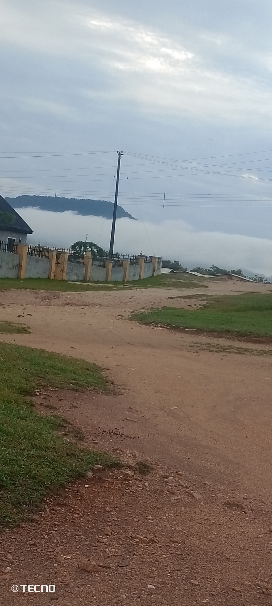 An overcast sky with scattered clouds looms over a rural landscape in Adankolo, Nigeria. In the foreground, a dirt road curves through patches of green grass. A low, grey concrete wall topped with a metal fence encloses a property with a house visible on the left. Beyond the wall, a dense layer of fog partially obscures a tree and a distant, mist-shrouded hillside. A solitary utility pole with wires stands near the fence. The scene appears to be captured in the early morning or late afternoon due to the muted lighting. The overall atmosphere is tranquil and somewhat ethereal due to the presence of the fog.