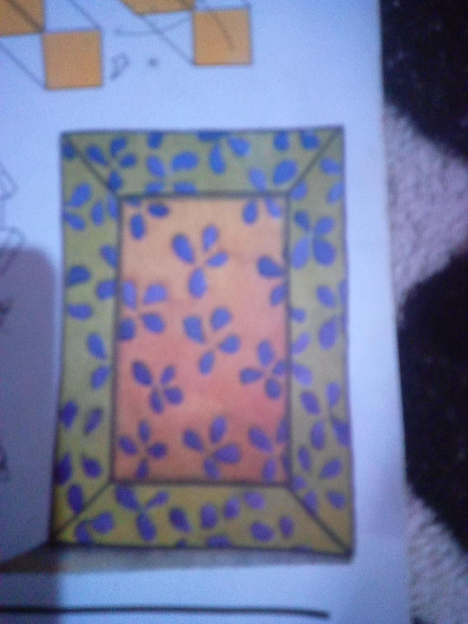 The image displays a close-up view of a drawing on a light-colored surface. The drawing features a rectangular frame with a patterned border. The border is a light green color adorned with scattered blue floral-like shapes. The central area of the frame is a peachy-orange color, also decorated with similar blue floral shapes. The drawing has a distinct border made of thin black lines, which outlines both the outer edge of the frame and the inner rectangle. The background appears to be a sheet of paper with some faint, abstract geometric shapes in the upper left corner, rendered in orange and white. The overall impression is that of a decorated picture frame design. There is no discernible information in the image that relates to the city of La Tagua, Venezuela.