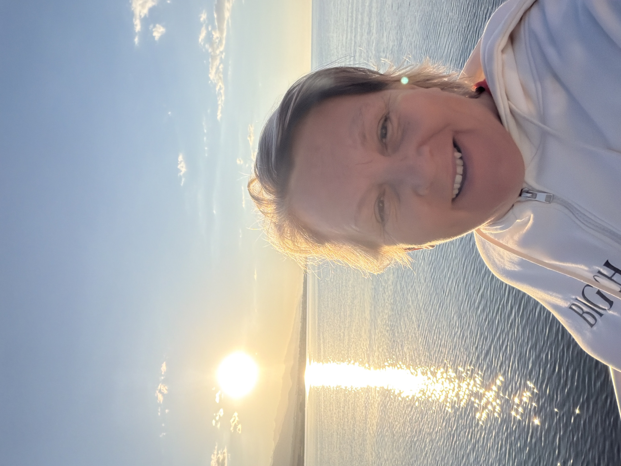 This is a selfie taken outdoors during daylight, likely sunset or sunrise, given the warm glow of the sun. The subject is a woman with short, light brown hair, smiling widely at the camera. She is wearing a white zippered hoodie, which has text on the sleeve that appears to read "BIG CH." The background consists of a large body of water, possibly a lake or ocean, reflecting the sun's light. The sky is a pale blue with a few wispy white clouds, and mountains are faintly visible in the distance. The water is rippling, suggesting a gentle breeze. The overall mood of the image is cheerful and relaxed, capturing a pleasant moment outdoors.