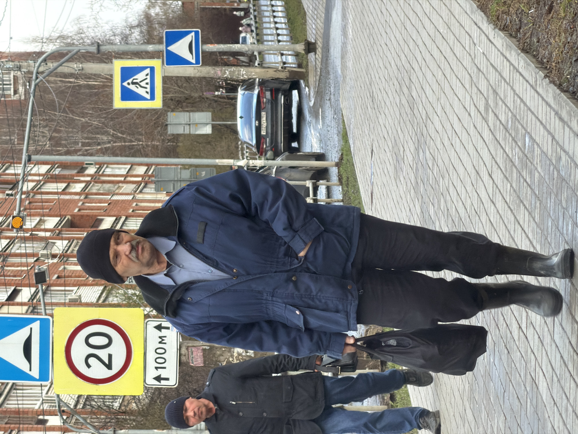 The image, set in Izhevsk, Russia, depicts an urban street scene on what appears to be a cool, overcast day, likely in late autumn, winter, or early spring, given the leafless trees and warm clothing. In the foreground, a man with a mustache, wearing a black beanie, a blue jacket, a light blue collared shirt, black pants, and black boots, looks towards the camera with a neutral to slightly amused expression. He holds a black bag in his left hand.
Behind him, another man in a black jacket and black beanie walks away from the camera. The scene features a tiled sidewalk to the right, bordered by a metal fence or guardrail. A road runs parallel to the sidewalk, where a black car is partially visible in the distance. Its Russian license plate, "X202XA 18", confirms the Udmurt Republic region, where Izhevsk is located. The pavement appears damp in areas.
Several road signs are prominent. These include a blue square sign with a white triangle and black bump, indicating an uneven road or speed bump; a yellow square sign with a blue pedestrian icon, marking a pedestrian crossing; and another blue square sign with a white triangle and black horn, possibly a warning about a sound signal ahead. Below these, a yellow square sign displays a '20' in a red circle, denoting a 20 km/h speed limit, next to a white rectangular sign with an upward arrow and '100M', indicating '100 meters ahead'. A traffic light with an illuminated yellow/orange signal is also visible. In the background, multistory red brick buildings with numerous windows and balconies line the street.