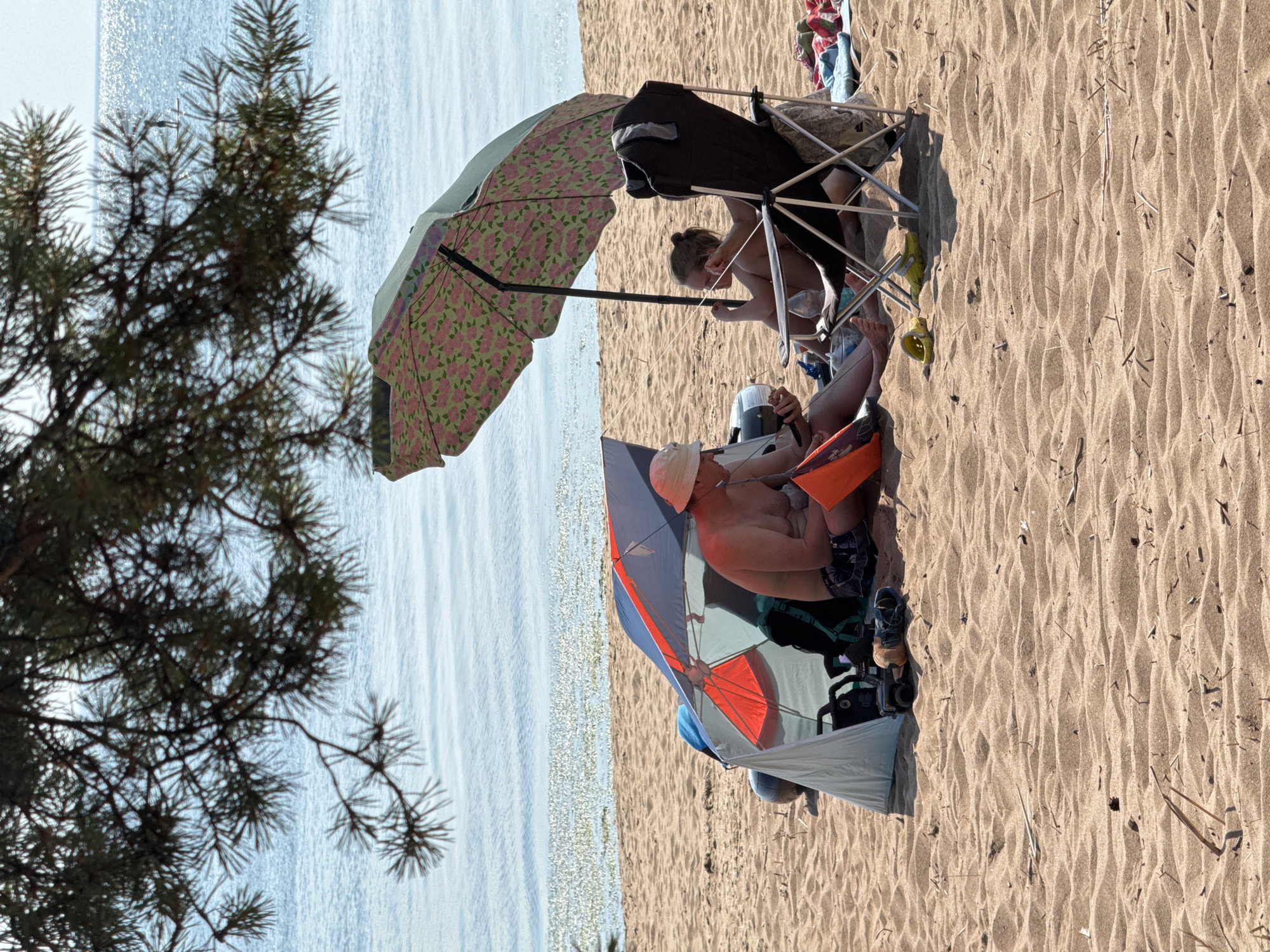 A wide shot captures a beach scene with two people seeking shade under umbrellas, with the Baltic Sea in the background. The sand is rippled with the texture of the beach. On the left, a man in a white hat and dark swim trunks sits on the sand under a beach tent. He is holding an orange object. To his right, a young person sits in a folding chair under a large, floral umbrella. They are looking to their left. A dark camping chair with a gray bag is next to them. In the background, the water of the sea is calm, with a slight sparkle from the sun. The branches of a pine tree hang in the upper portion of the frame, partially obscuring the sky.