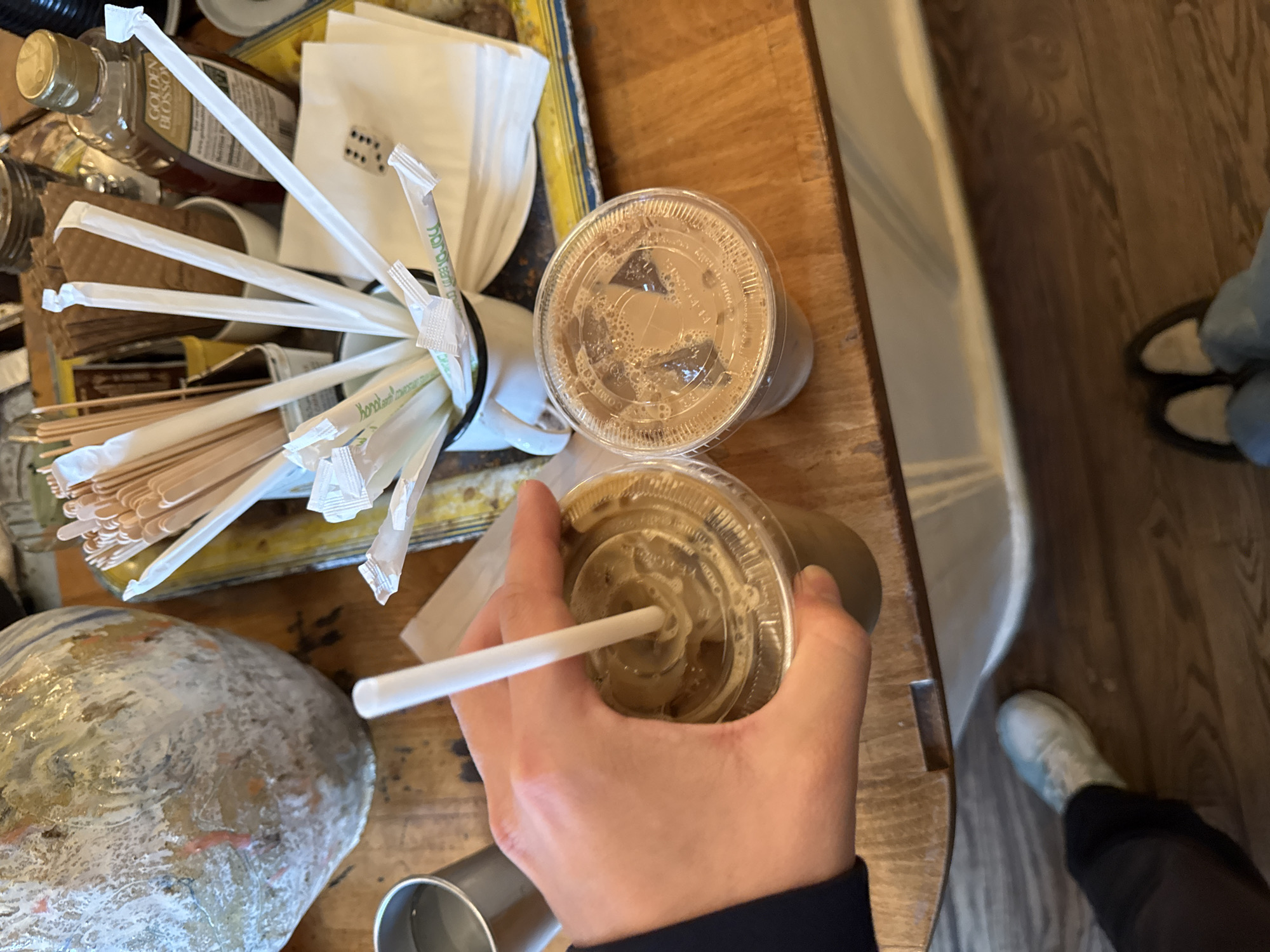 The image shows a wooden table, possibly in a coffee shop. The table is cluttered with various items, including a container filled with straws, a jar of honey, napkins, and two iced coffee cups with lids. One iced coffee cup is being held in a hand, with a straw inserted. In the background, partially visible, are sections of the floor and lower leg and feet of people sitting nearby. Based on the presence of "Golden Blossom" honey, a product widely available in the United States, it is very likely that this image was taken somewhere in the United States. I have no information to identify a location of this type. I cannot provide the specific location in the city of New York, United States of America.