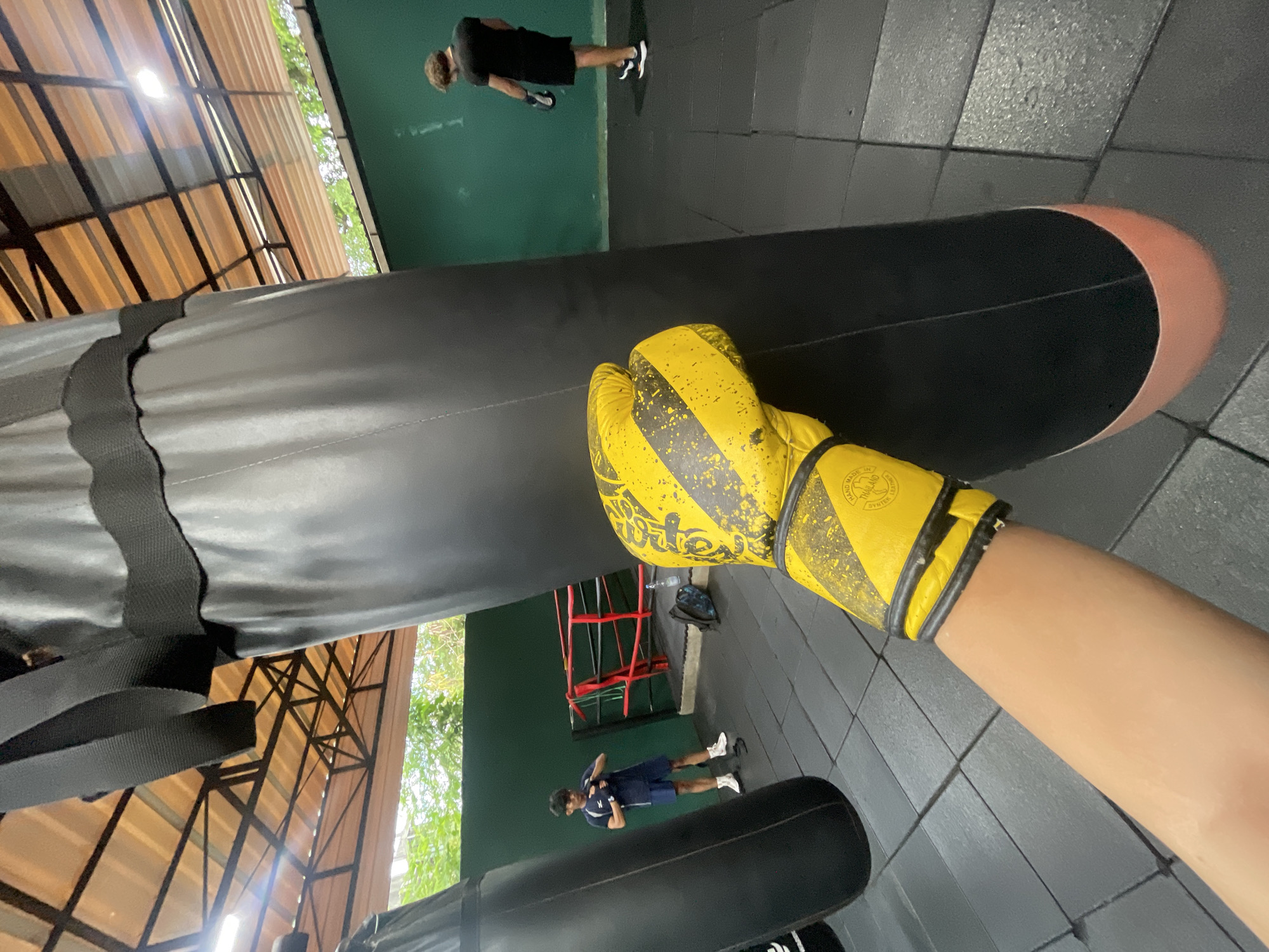 The image depicts the interior of a boxing gym, likely in Kuta Selatan, Indonesia, given the lighting and tropical foliage visible through an open roof. In the foreground, a person's arm, adorned with a yellow and black boxing glove, is extended towards a large black punching bag. The glove is speckled with wear and tear. Behind the punching bag, another individual wearing a black t-shirt and shorts is seen facing away from the camera, with boxing gloves on. To the left, a second person, dressed in a dark blue t-shirt and shorts, stands near a boxing ring, appearing to be adjusting something on their hands. The gym floor is covered in black rubber mats. The space has an open-air structure with a corrugated metal roof, allowing natural light to enter. The walls are painted a deep green.