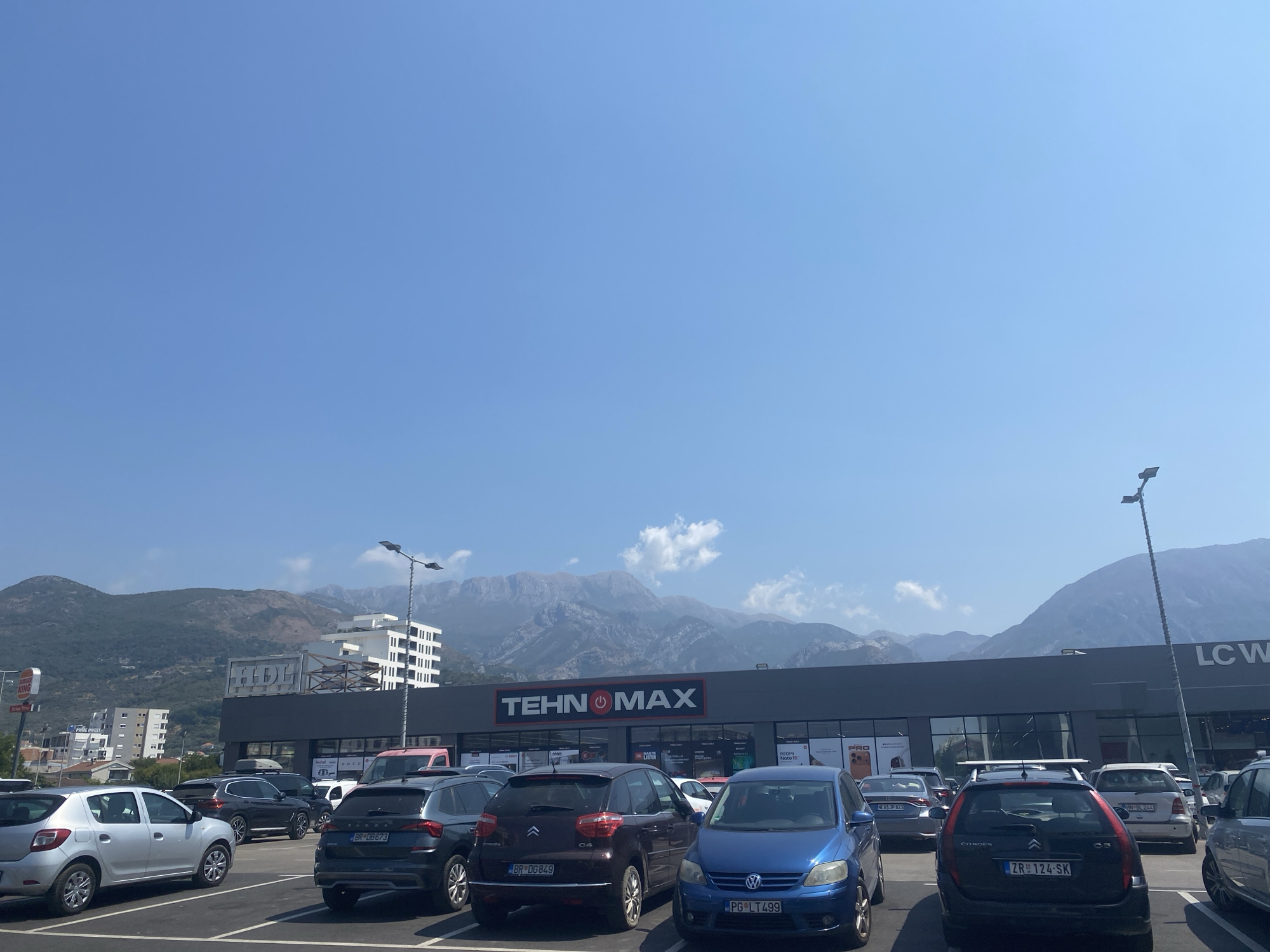 The image shows a parking lot in front of a retail store labeled "TEHNOMAX" and "LC WAIKIKI" in Bjeliši, Montenegro. The sky is clear and blue, with mountains in the background. Several cars are parked in the lot, including a silver Dacia Sandero, a dark blue Volkswagen Touran, a dark red Citroën C4 Picasso, and a dark grey Škoda Karoq. The TEHNOMAX store is a long, dark building with a large sign. The LC WAIKIKI store is located to the right of TEHNOMAX. There are no people visible in the image. The overall scene is one of a typical commercial area on a sunny day.