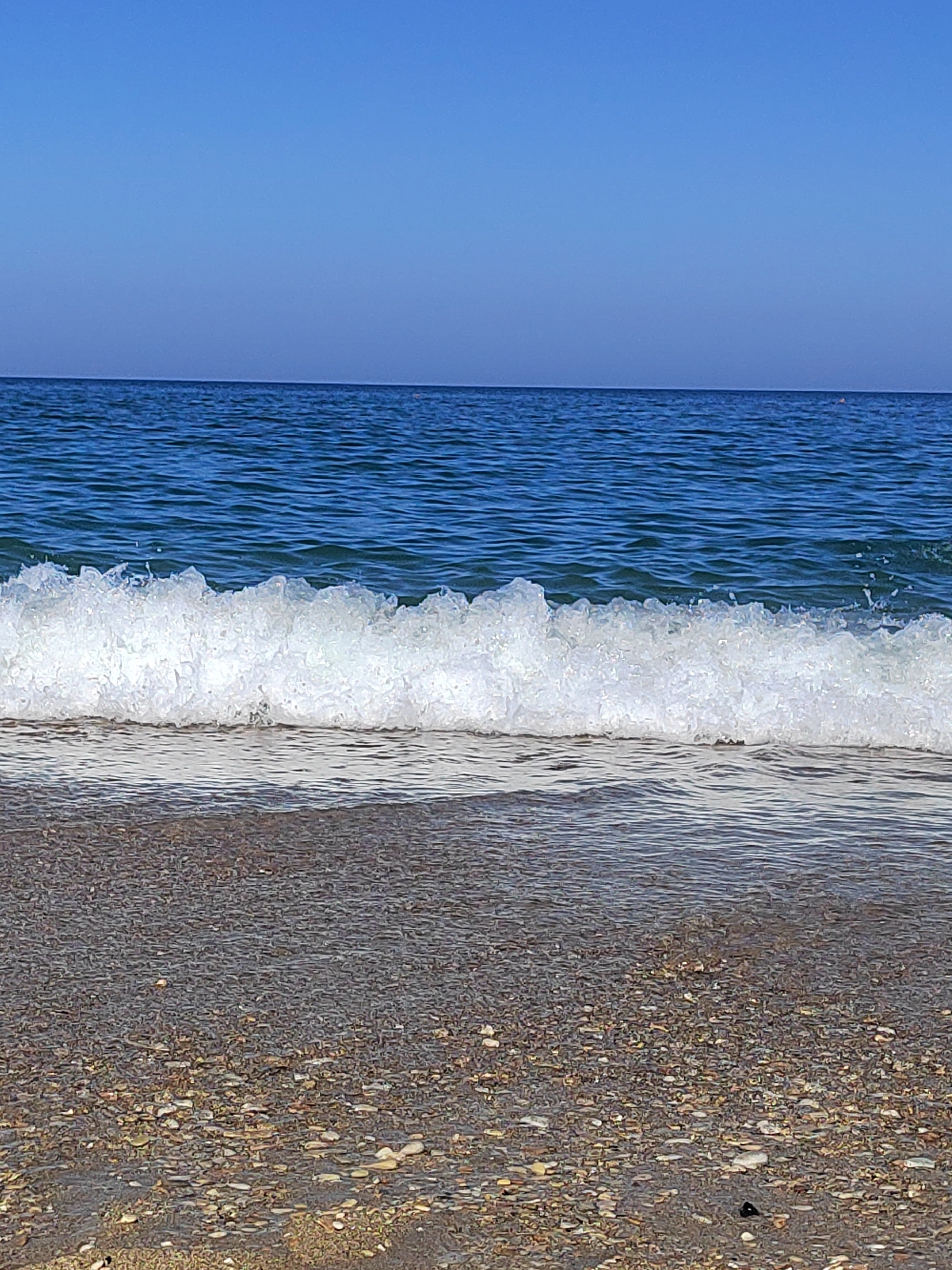 A close-up, eye-level, vertical medium shot captures a calm sea meeting a pebbled beach in Porto Recanati, Italy. The sky, a clear, gradient blue, occupies the top third of the frame. Below it, the deep blue sea stretches horizontally, with a gentle wave cresting in the mid-ground, its white foam glistening. The foreground is dominated by the textured surface of the beach, composed of numerous small, smooth pebbles of varying shades of brown, grey, and white. The water gently laps at the shore, creating a transparent layer over some of the pebbles. The scene is devoid of people or any man-made objects, emphasizing the natural beauty of the coastal environment.