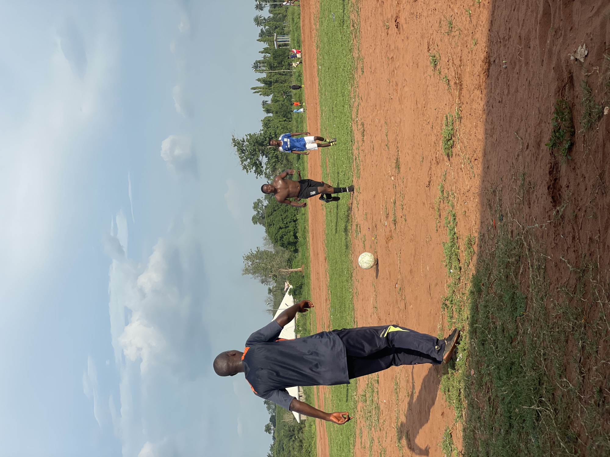 The image depicts an outdoor scene on what appears to be a soccer (football) field, captured with a 90-degree clockwise rotation, positioning the sky on the left and the ground on the right.
In the bottom-left foreground, a dark-skinned man is seen from behind, wearing a dark blue and orange short-sleeved top and dark blue track pants with yellow accents. The brand "UHLSPORT" is visible on his pants. He is in a dynamic pose, suggesting movement or preparation for an action. A white soccer ball rests on the reddish-brown ground near him.
Further to the right in the midground, two other dark-skinned men are visible. One is shirtless, wearing black shorts with the "NIKE" logo written in white on his left thigh. He is captured mid-stride, seemingly running or jogging, with his body turned slightly to the left. Behind him, another man in a blue and white short-sleeved shirt and white shorts stands upright, looking towards the right. Other individuals are faintly visible in the far background.
The playing surface is characterized by a mix of reddish-brown dirt and patches of green grass. The background (top-right of the image) features a line of trees, including distinctive palm trees, and some distant white structures. The sky on the left side of the image is light blue with scattered white, puffy clouds, indicating bright daylight, likely late morning or early afternoon, with clear and warm weather. The visible shadows suggest a strong sun.
The activity taking place appears to be a casual soccer game or practice, given the players' attire, dynamic poses, and the presence of a ball on the field. The scene offers a glimpse into outdoor sports in Ogbogbo, Nigeria.